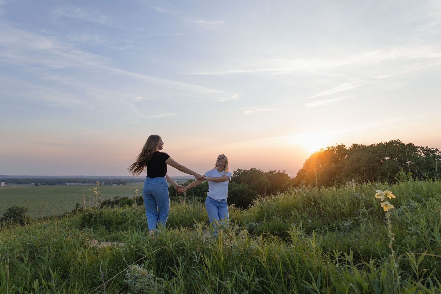 Sioux City, Iowa photographer. Two young women holding hands walking through tall grass at sunset with scenic countryside views.