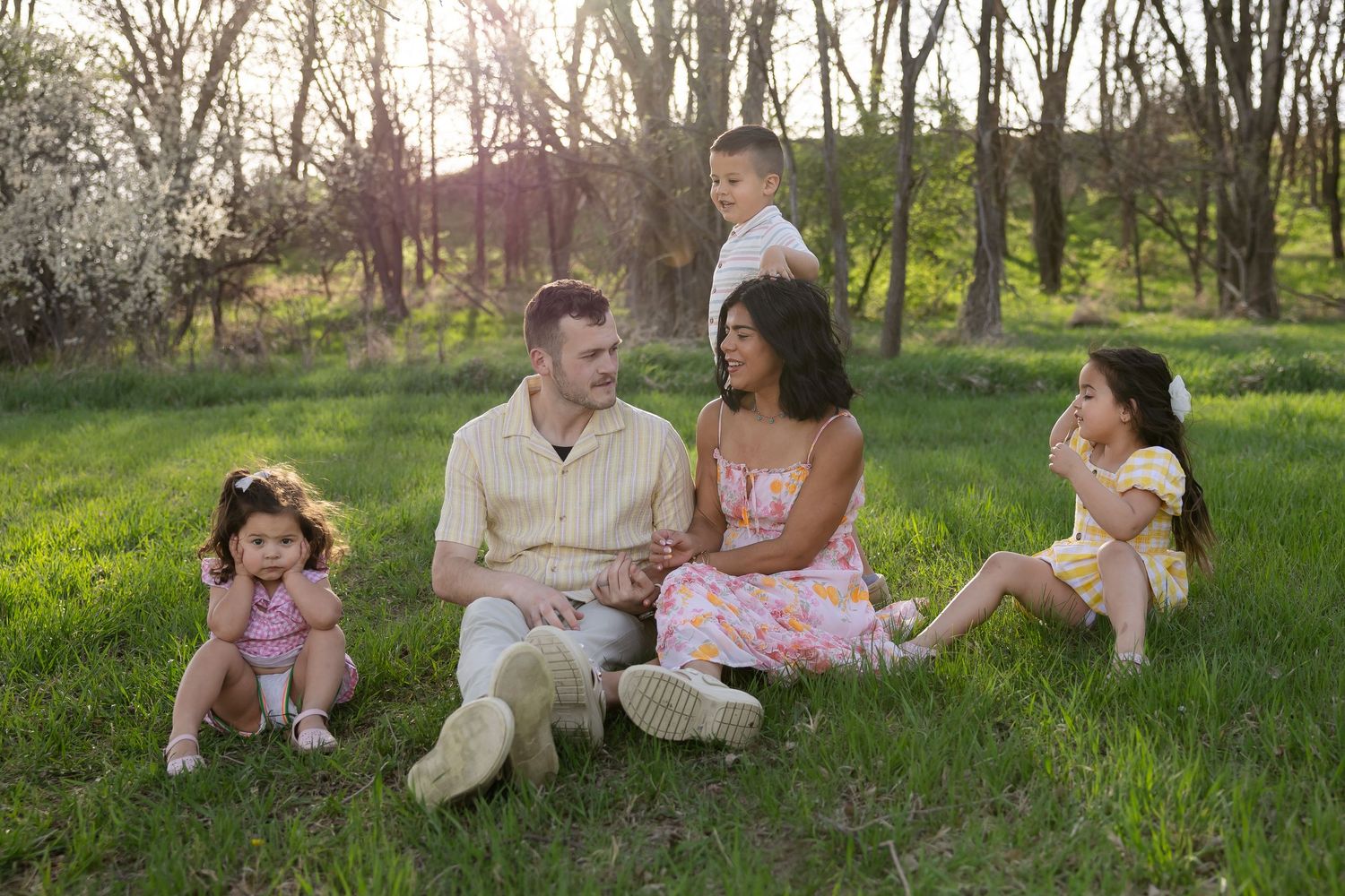 Sioux City, Iowa photographer. A happy interracial family with three young children sits together on green grass in a sunlit spring park.
