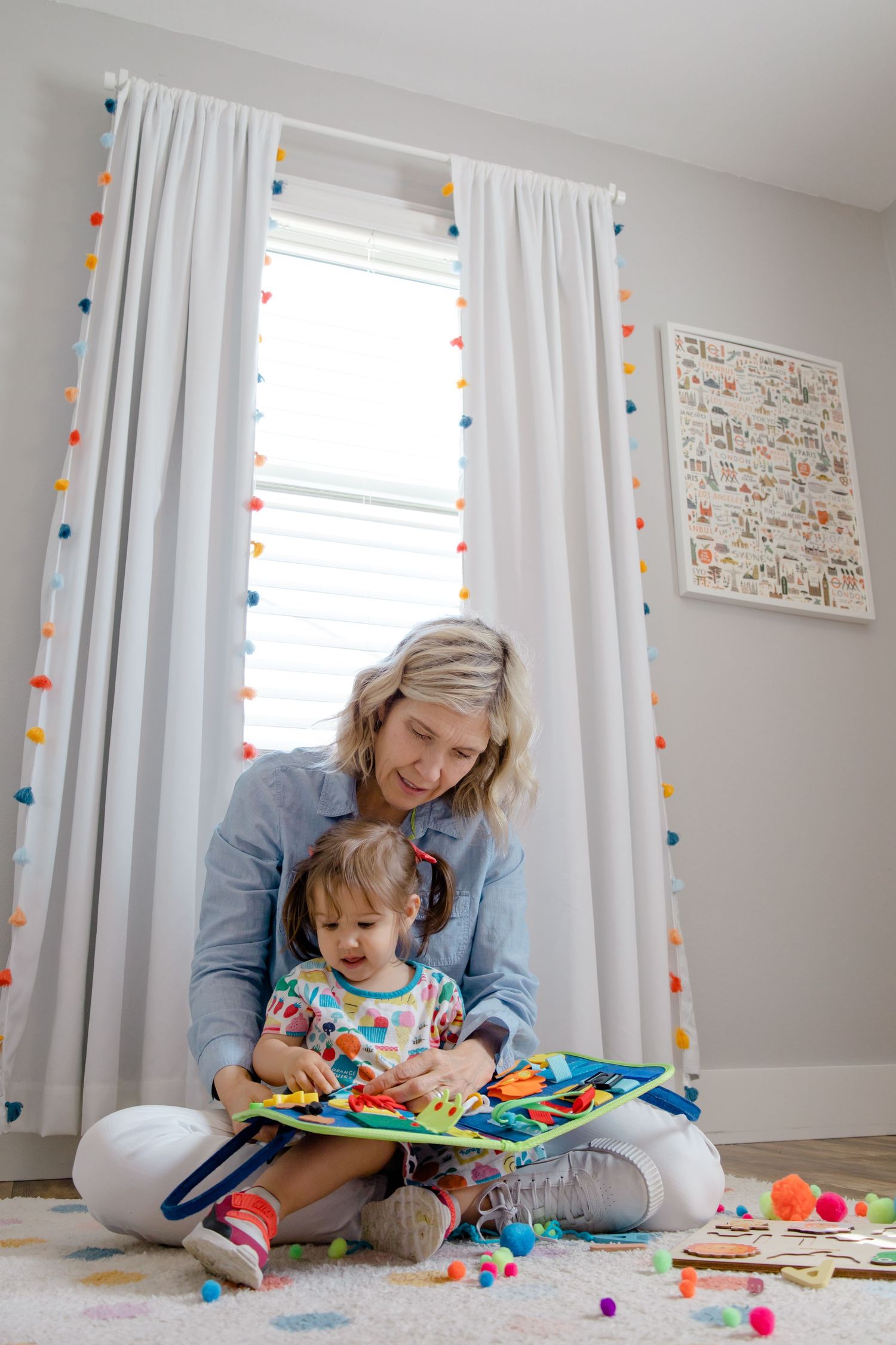 Sioux City, Iowa photographer. Blonde woman sitting cross-legged on floor reading colorful book with toddler in bright nursery room.
