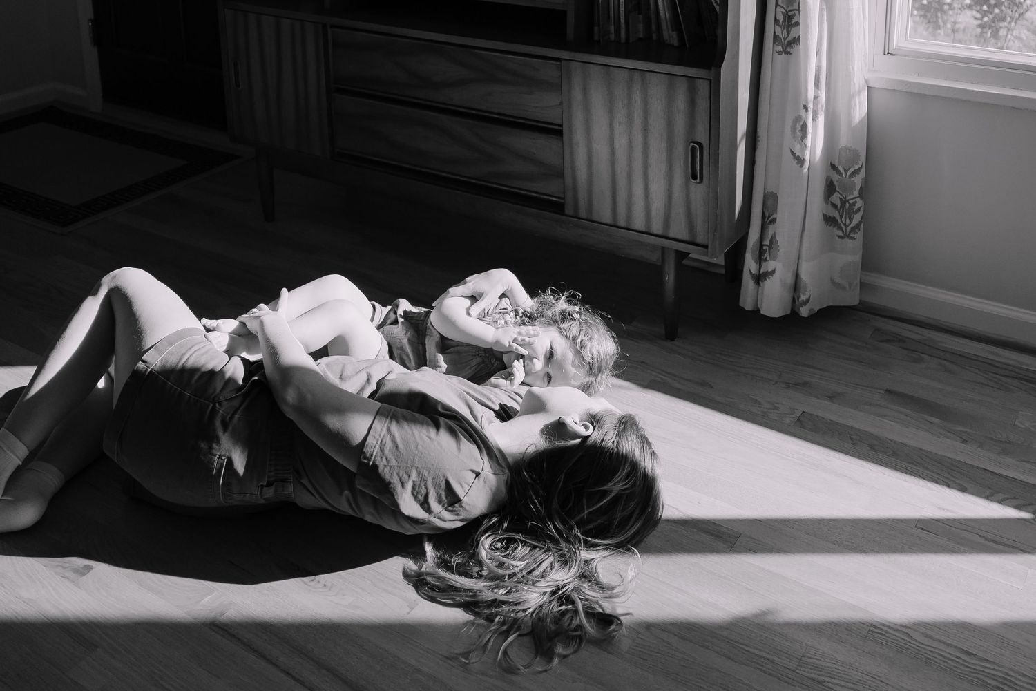 Sioux City, Iowa photographer. Black and white photo of a mother and child playing together on a sunlit hardwood floor indoors.