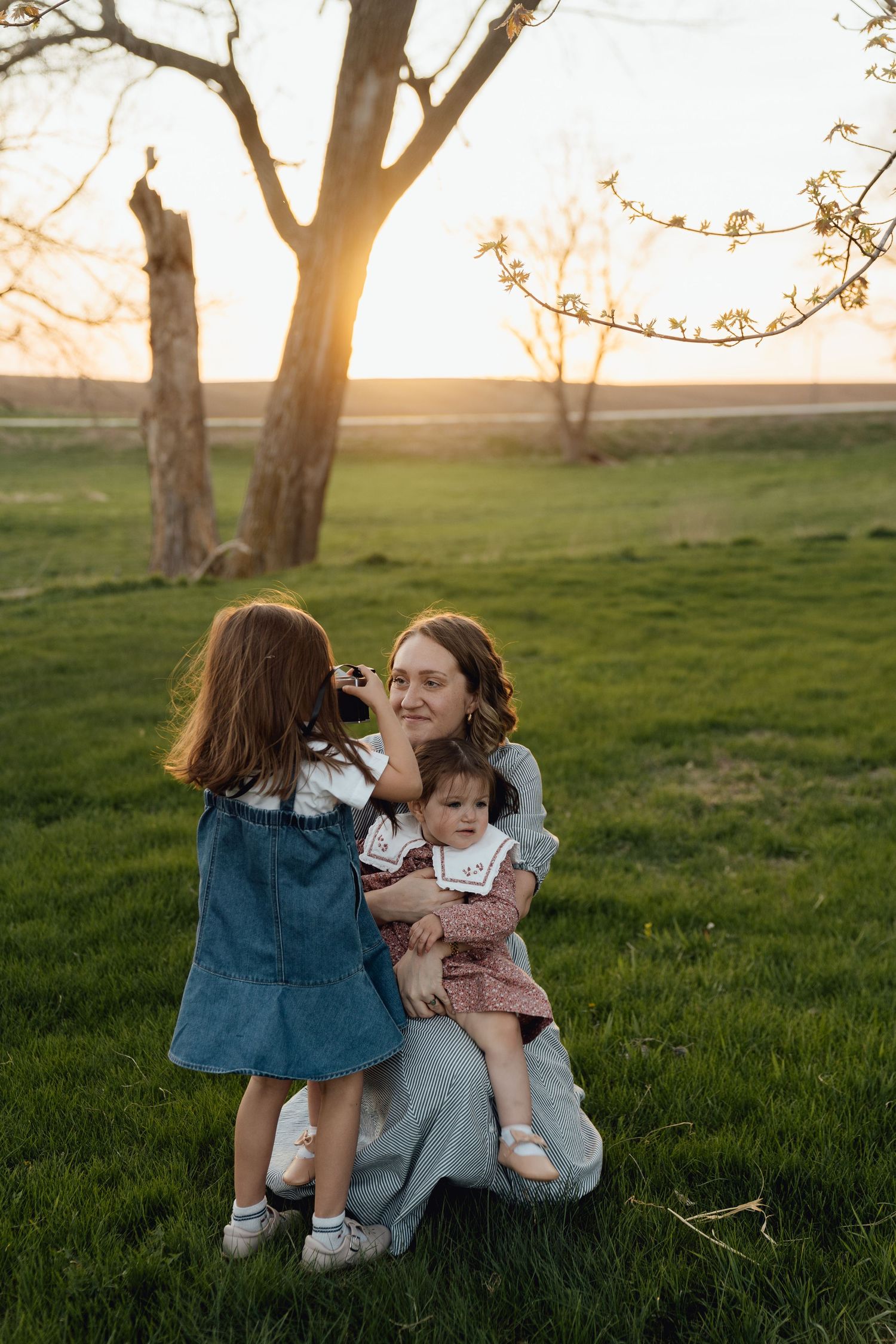 Sioux City, Iowa photographer. A mother kneels on green grass at sunset holding her baby while her young daughter touches her face.