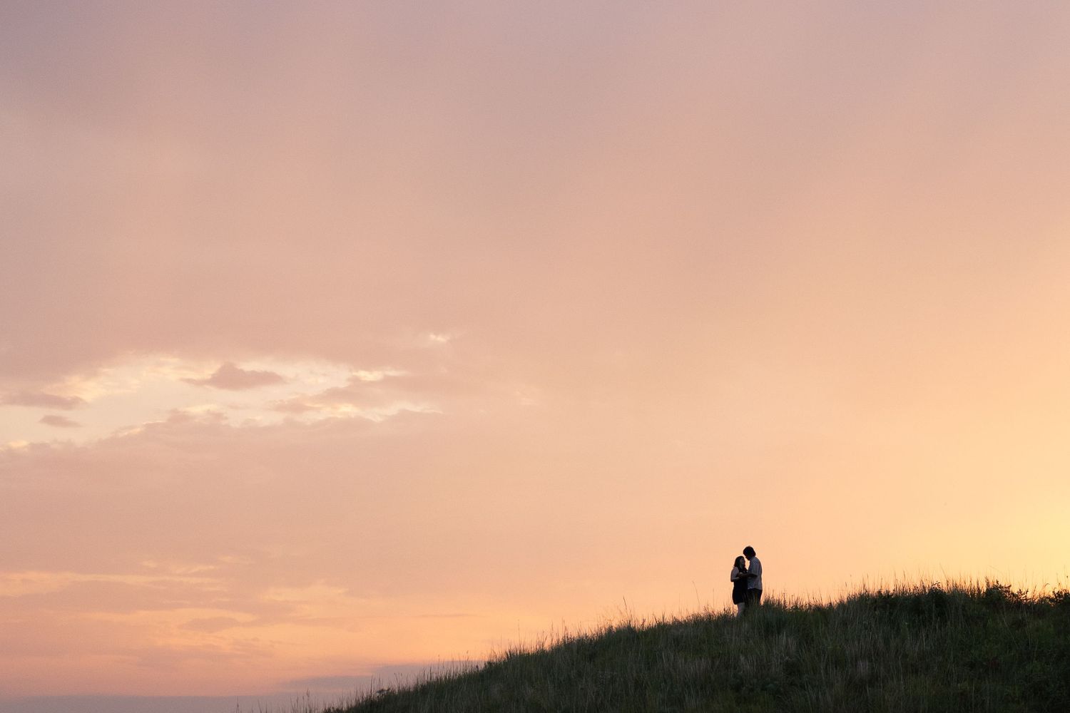 Silhouette of a couple embracing on a grassy hill at sunset with a pink and orange sky.