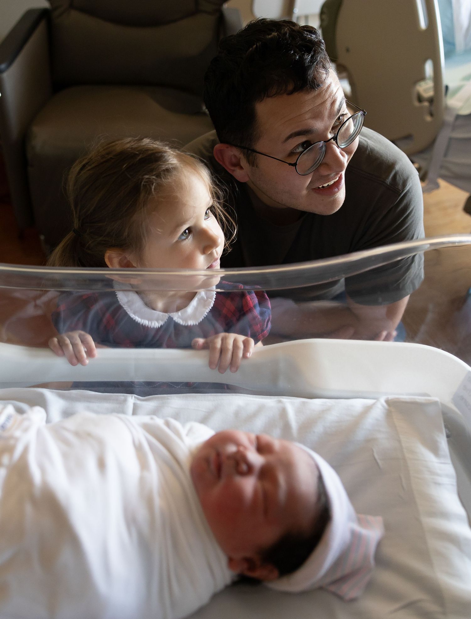 A man with glasses and young girl smile at a swaddled newborn baby in a hospital bassinet.
