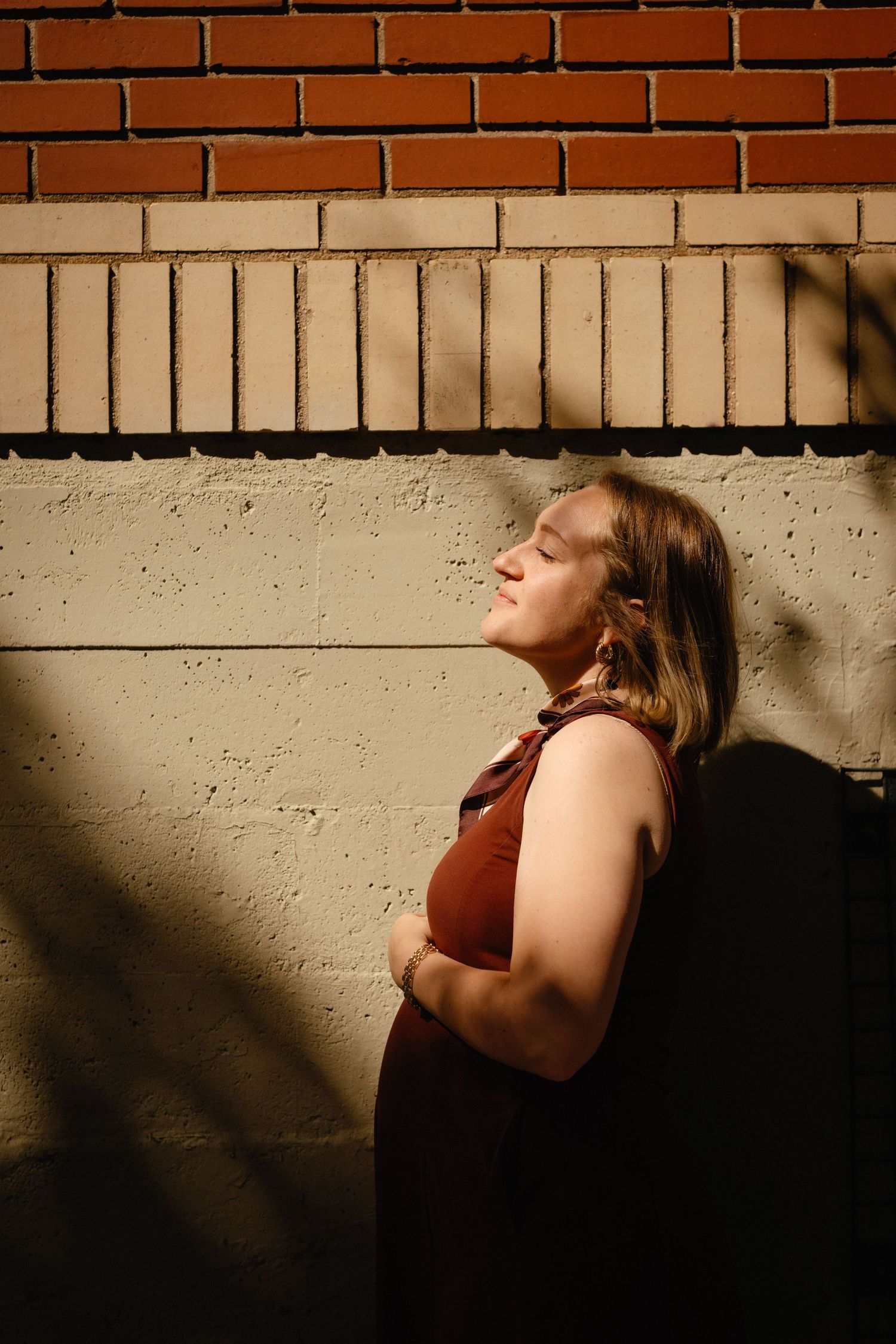 Woman in rust top leaning against brick wall with eyes closed, bathed in warm sunlight and shadows.