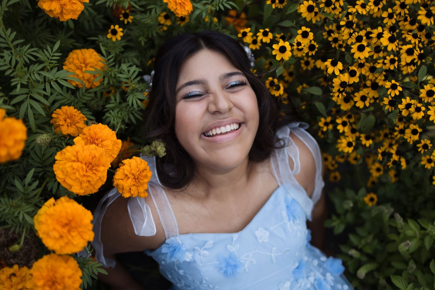 Young woman in blue dress smiling among vibrant orange marigolds and yellow flowers in a garden.