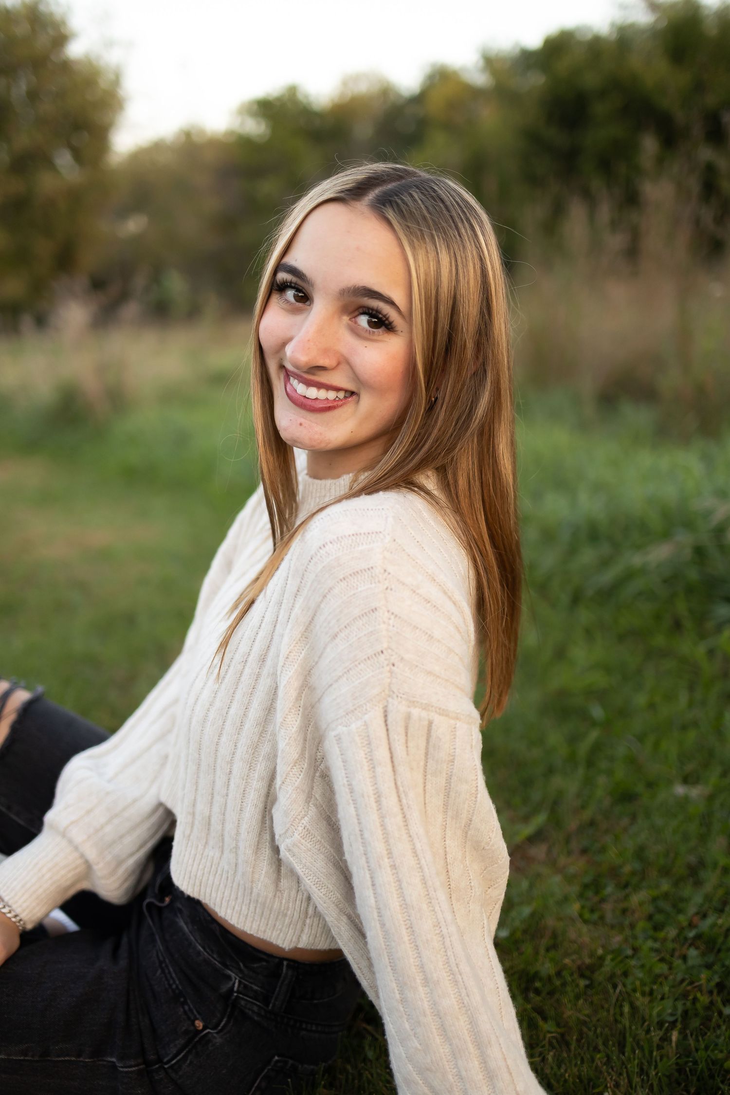 Sioux City, Iowa photographer. Smiling teenage girl with long blonde hair sitting in grass wearing a cream knit sweater during golden hour.