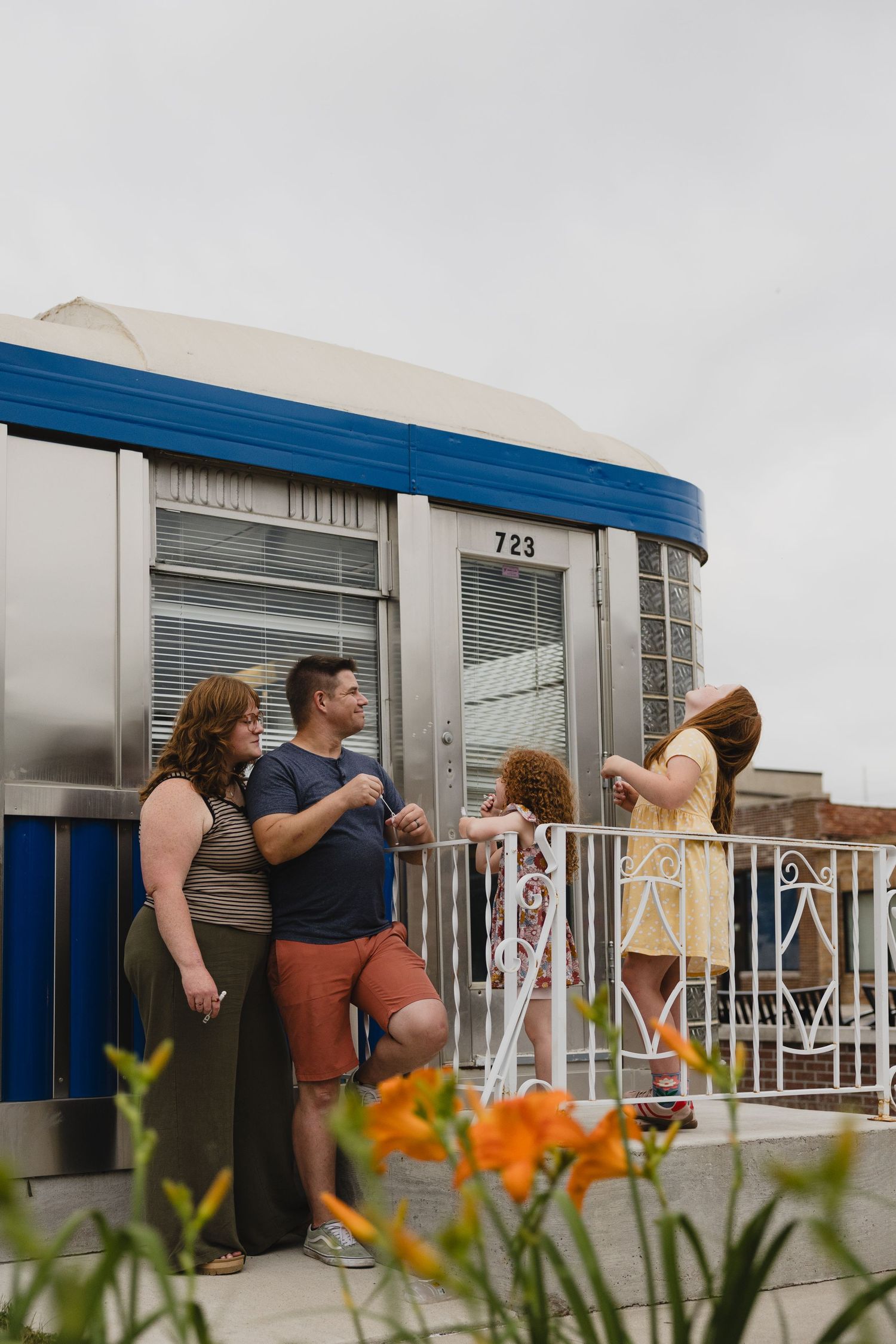 A couple and two children stand on a balcony outside a retro diner-style building numbered 723 with blue trim.