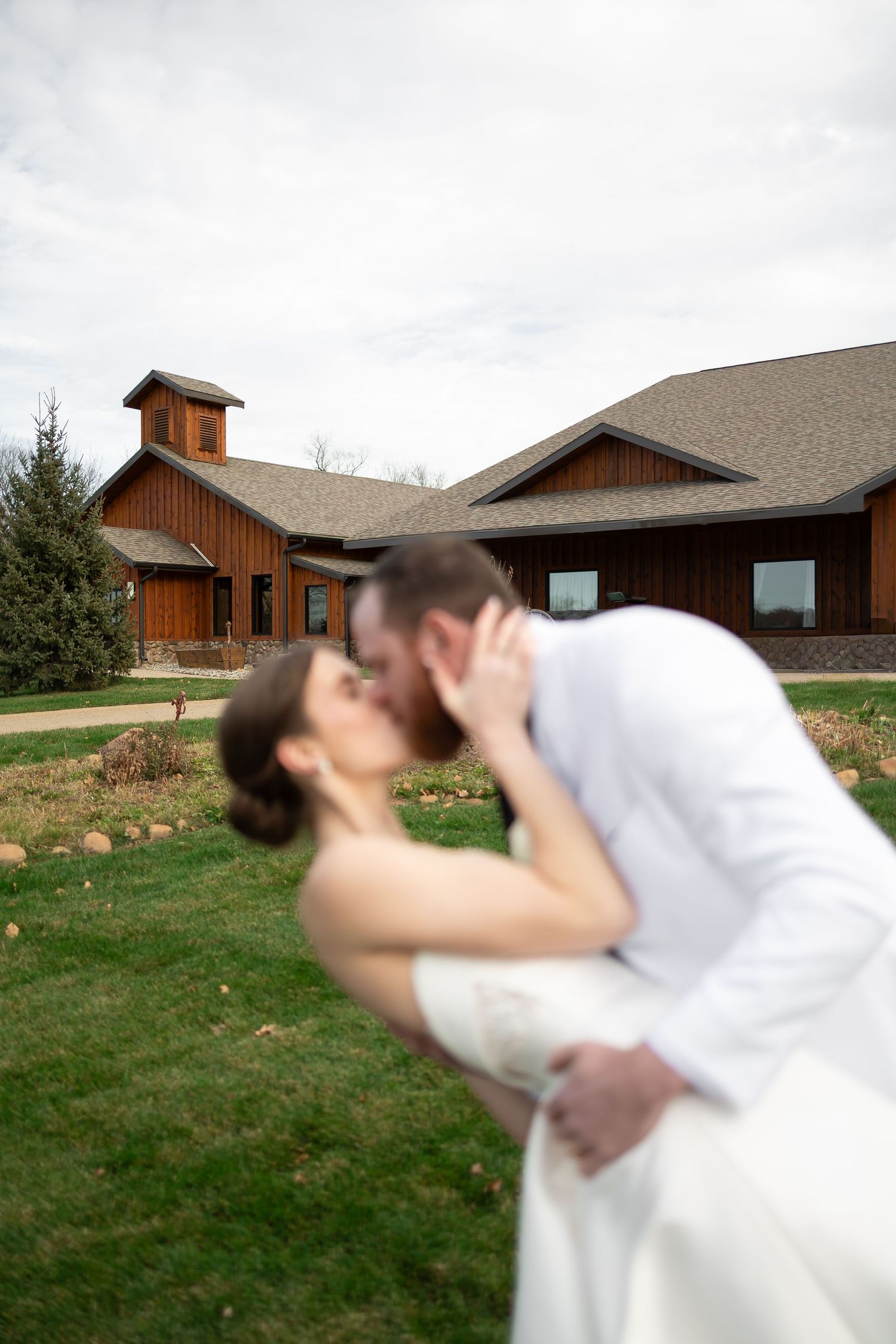 Sioux City, Iowa photographer. A bride and groom share a romantic dip kiss on a green lawn in front of a rustic wooden barn wedding venue.