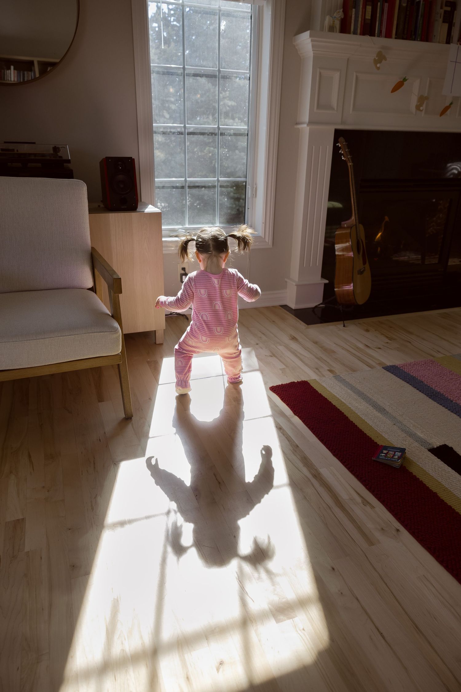 Sioux City, Iowa photographer. A toddler girl in pink pajamas walks toward a sunlit window casting a shadow on hardwood floors.