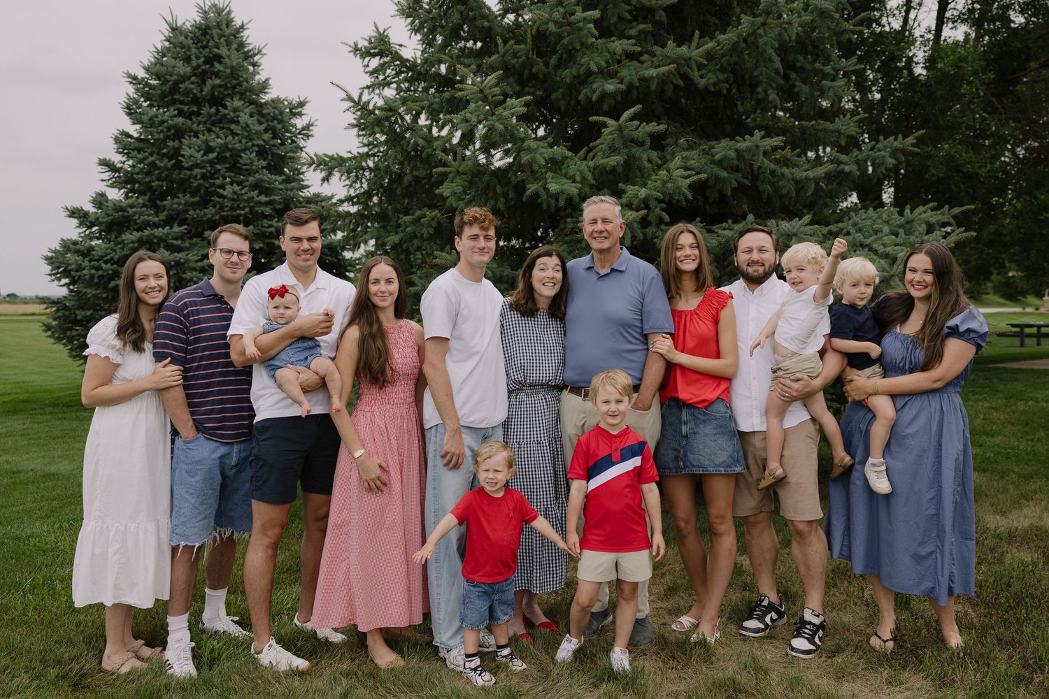Large multigenerational family posing outdoors on green lawn with evergreen trees, wearing red, white, and blue outfits.
