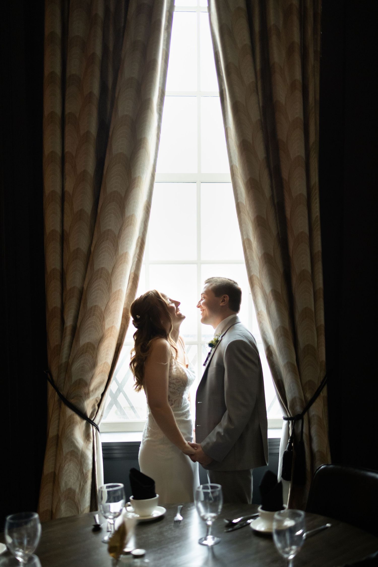 Bride and groom holding hands by a sunlit window with elegant drapes in a romantic wedding venue.