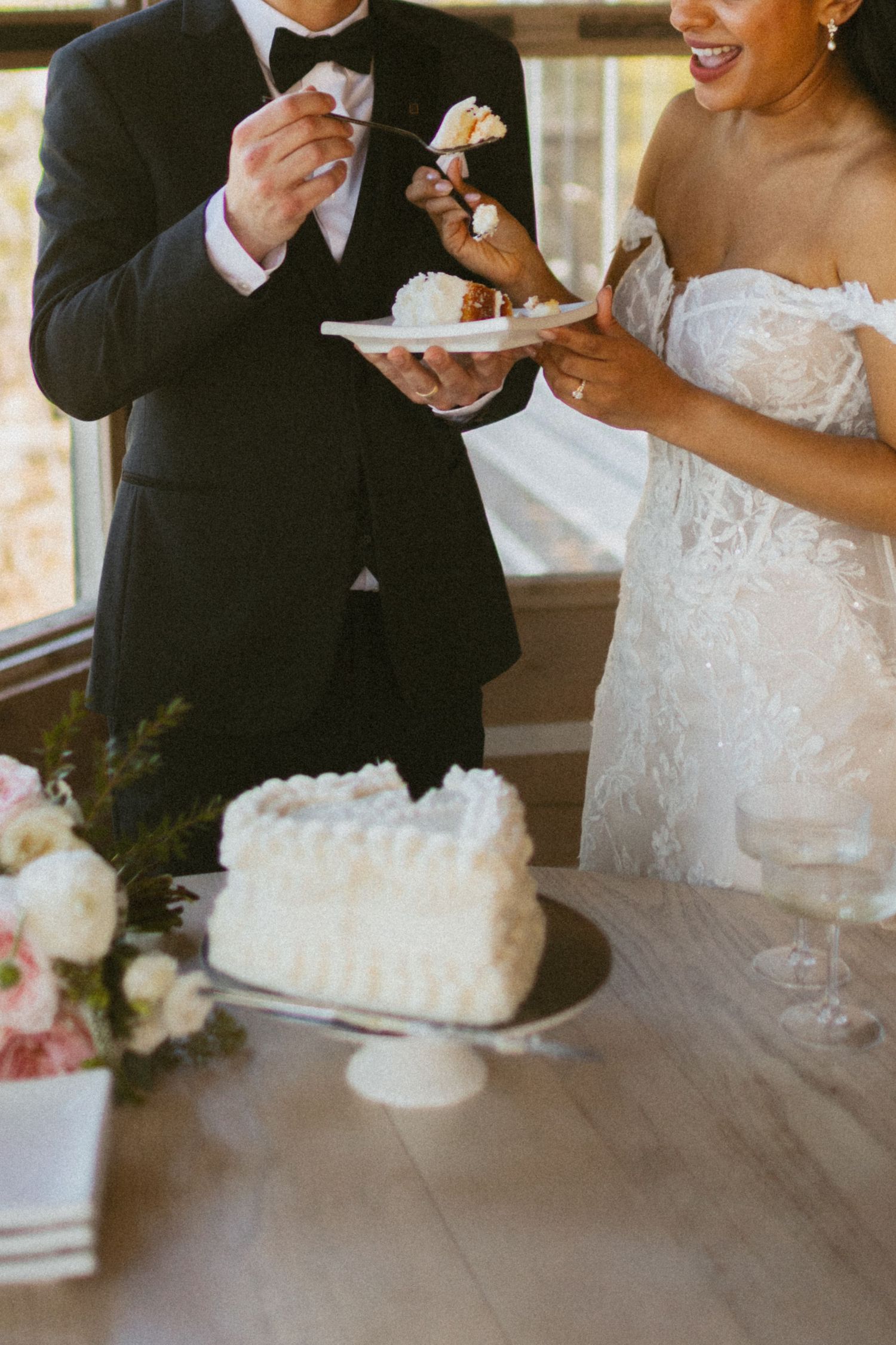 Bride and groom cutting and tasting a white heart-shaped wedding cake on a cake stand at their reception.