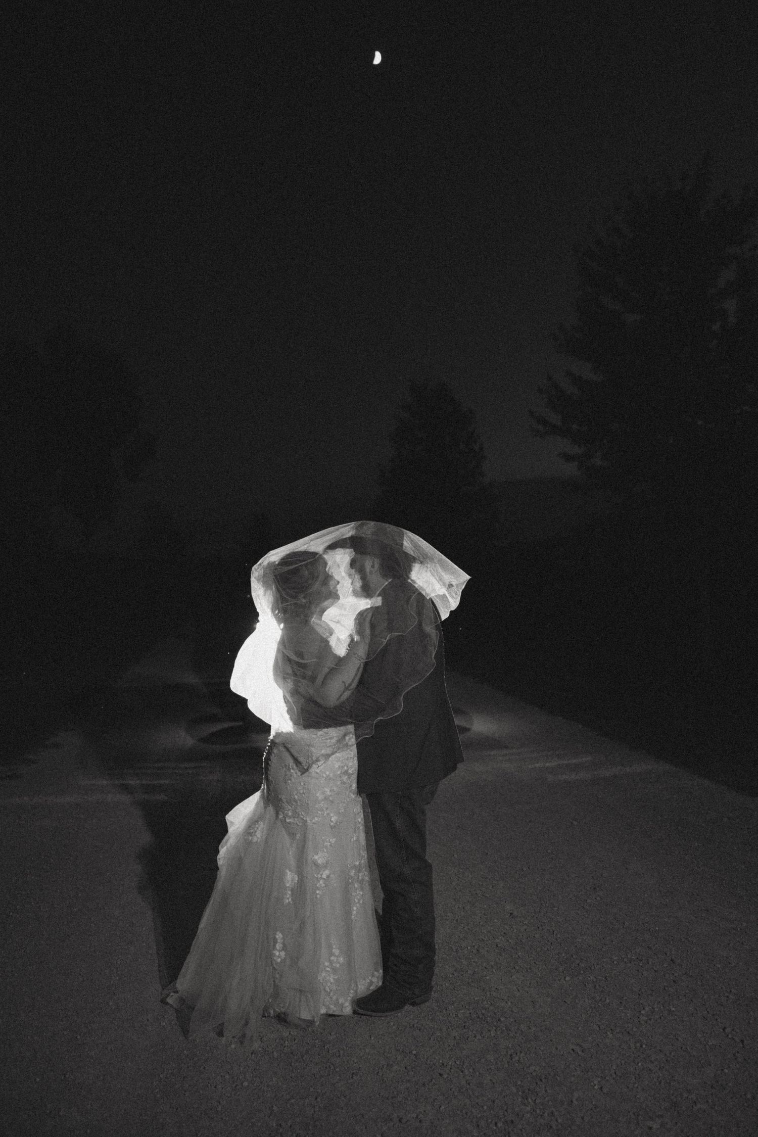 Black and white photo of bride and groom kissing under veil at night with crescent moon above.