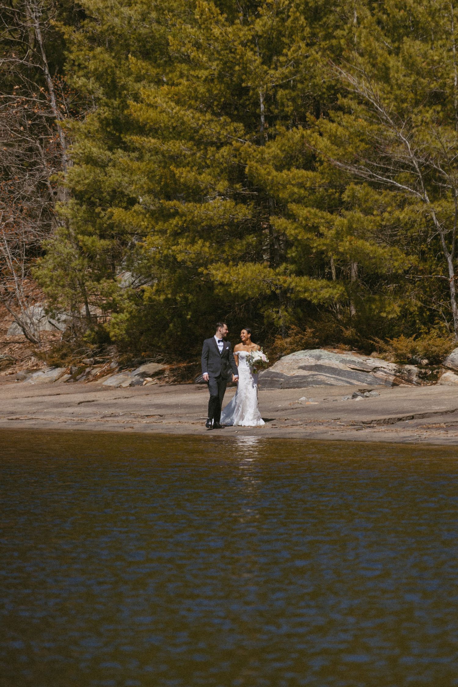 Bride and groom walk hand in hand on rocky lakeside shore surrounded by lush evergreen forest.
