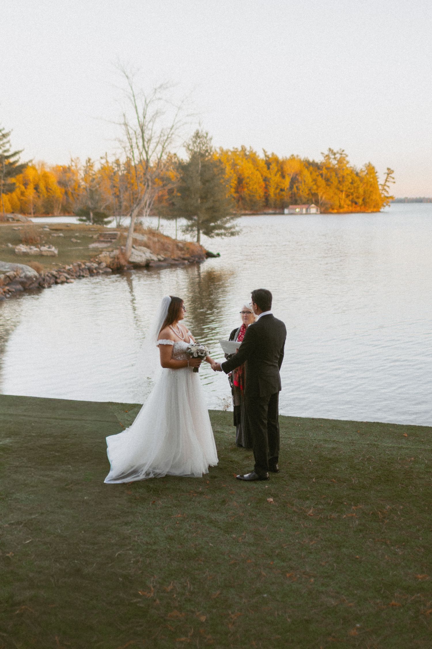 Bride and groom exchange vows lakeside with autumn foliage backdrop during outdoor wedding ceremony.