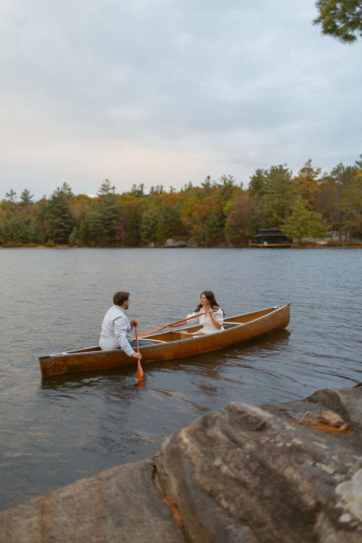 A couple paddling in a wooden canoe on a serene lake surrounded by autumn trees and rocky shoreline at dusk.