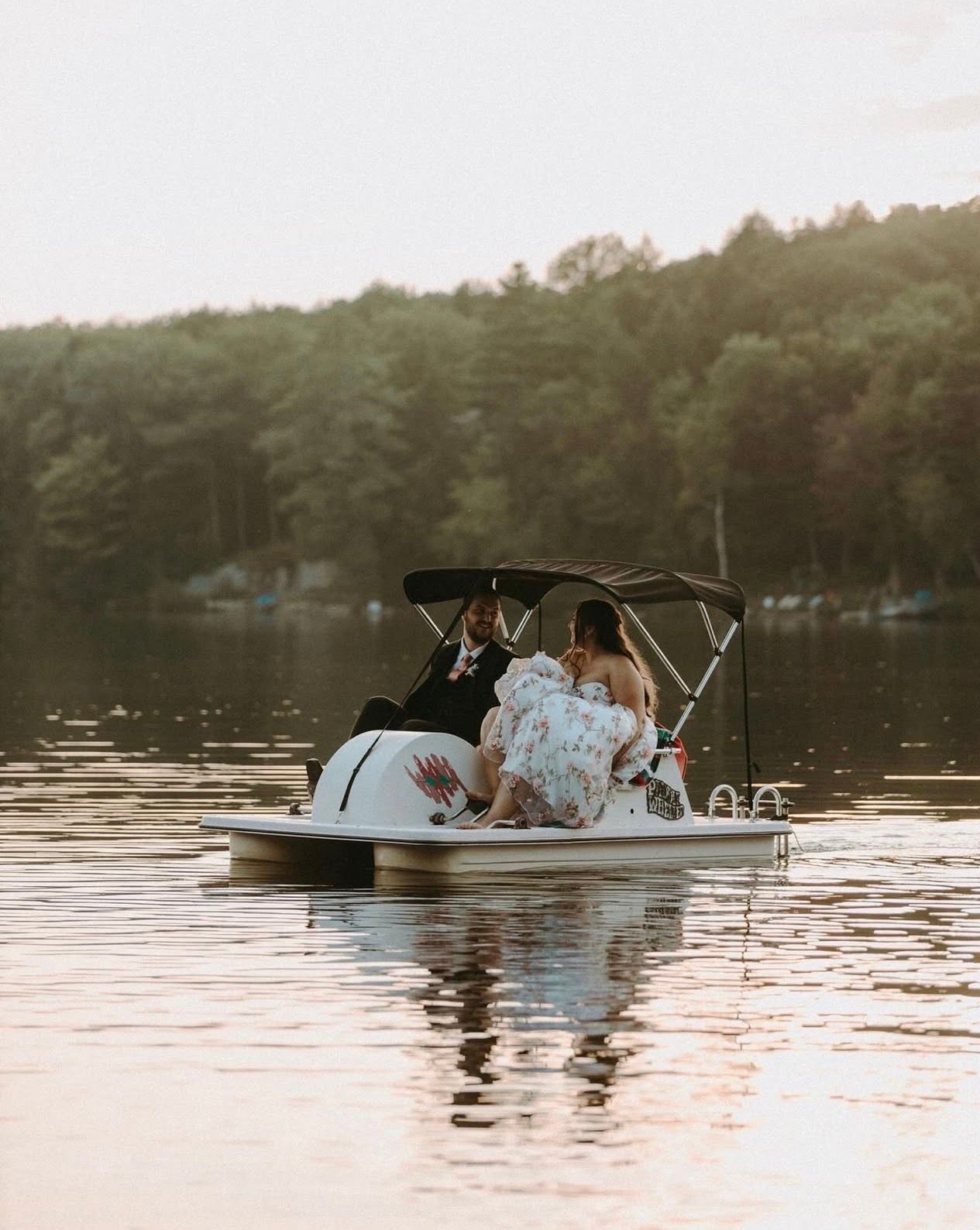 Bride and groom riding a swan paddle boat on a calm lake at sunset with lush green trees in the background.