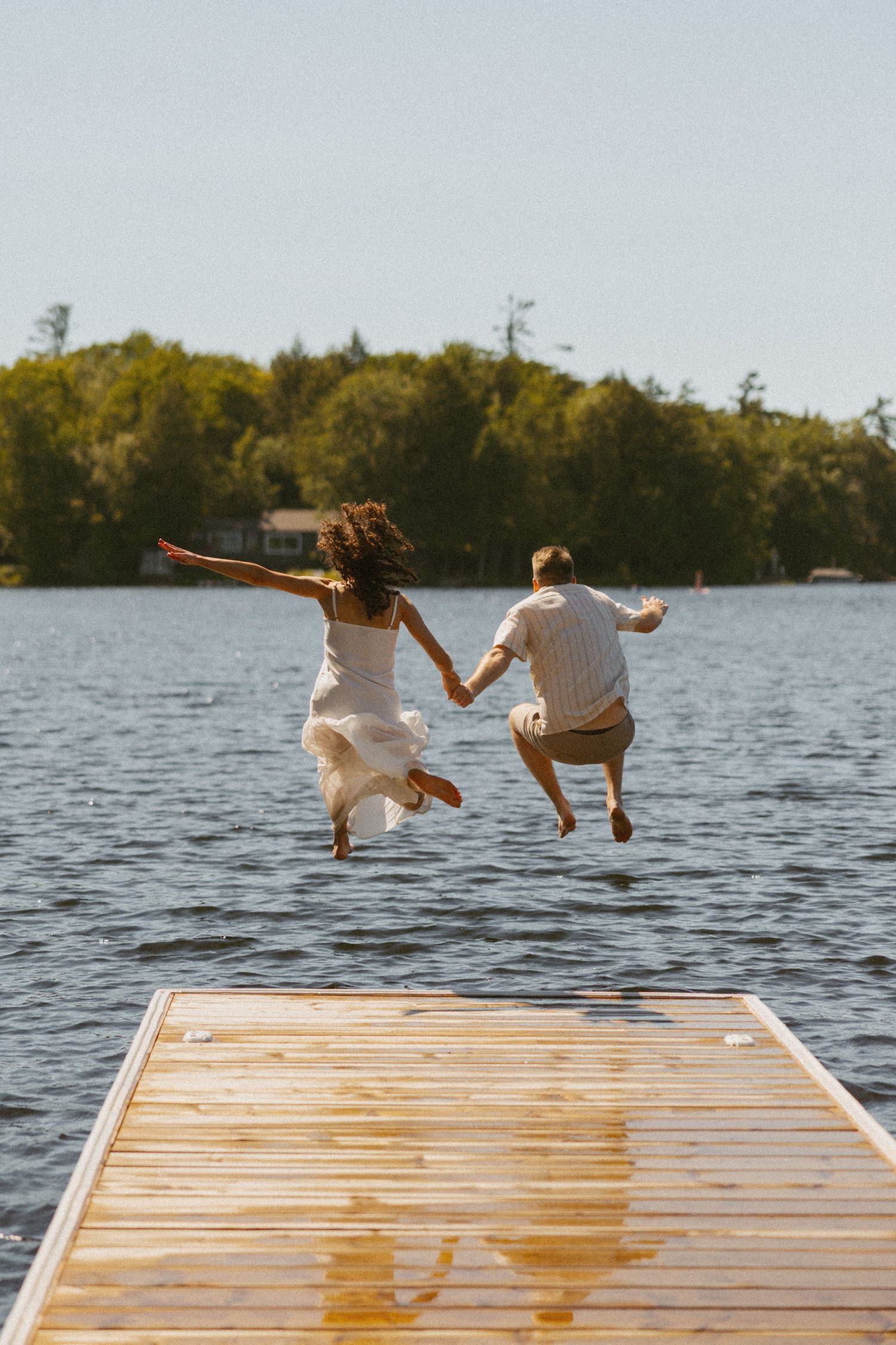 Couple holding hands jumping off a wooden dock into a lake, surrounded by lush green trees on a sunny day.
