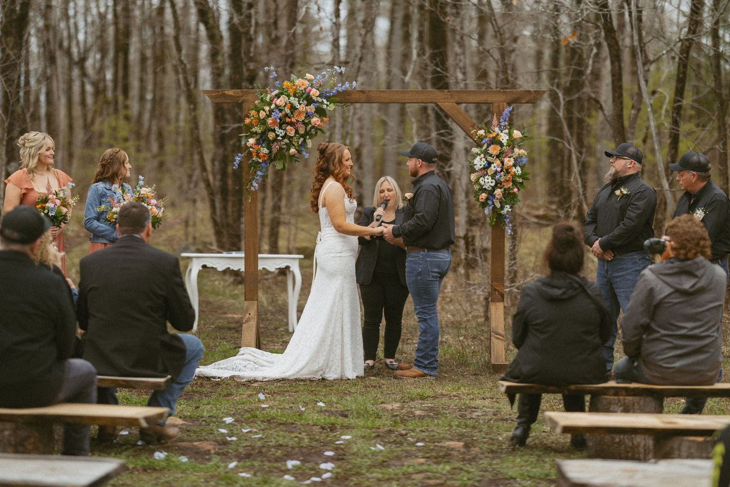 Outdoor woodland wedding ceremony with couple at floral arch, officiant, and guests seated on rustic benches.