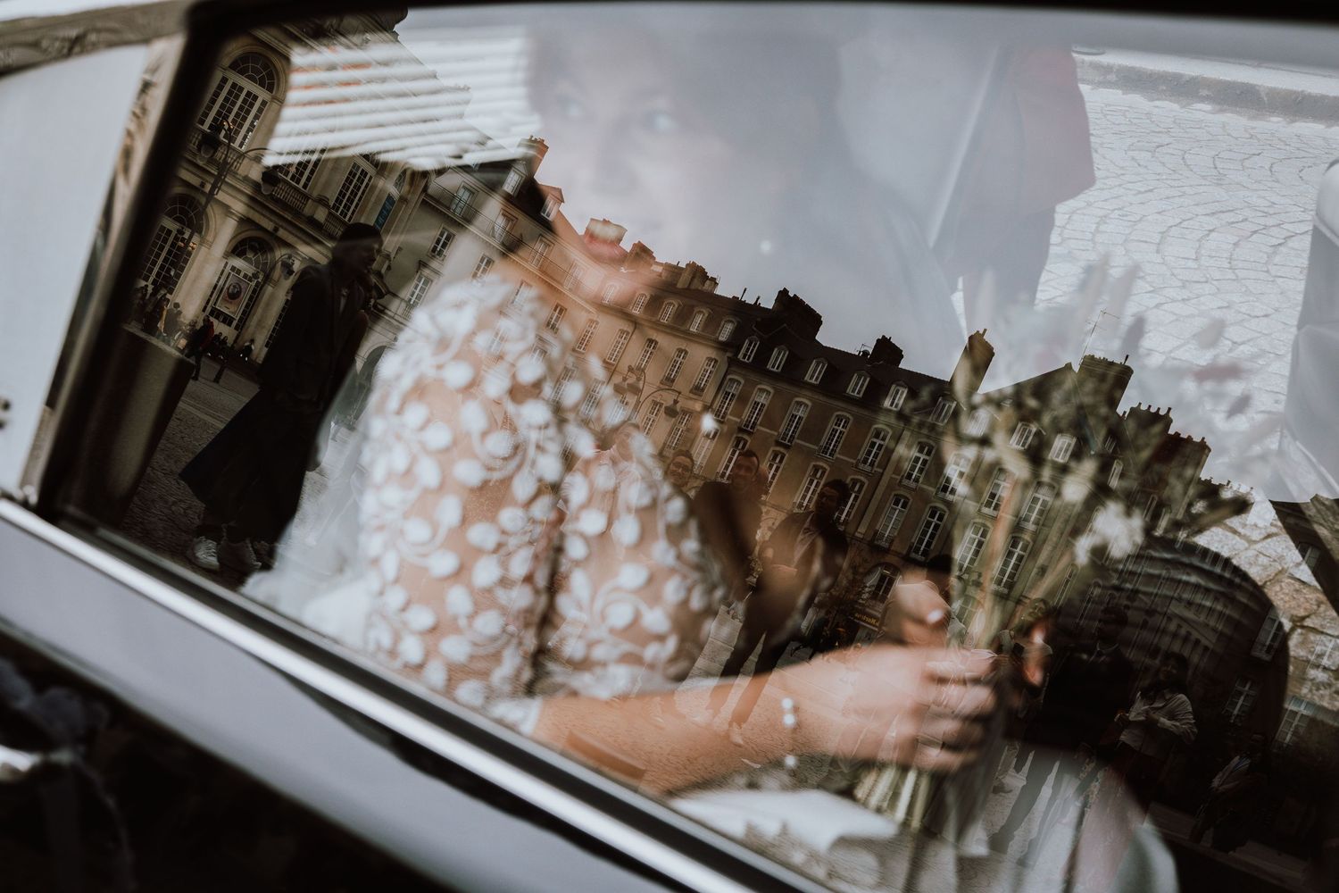 Through a car window reflection, someone in a white lace dress sits against blurred city architecture in the background.
