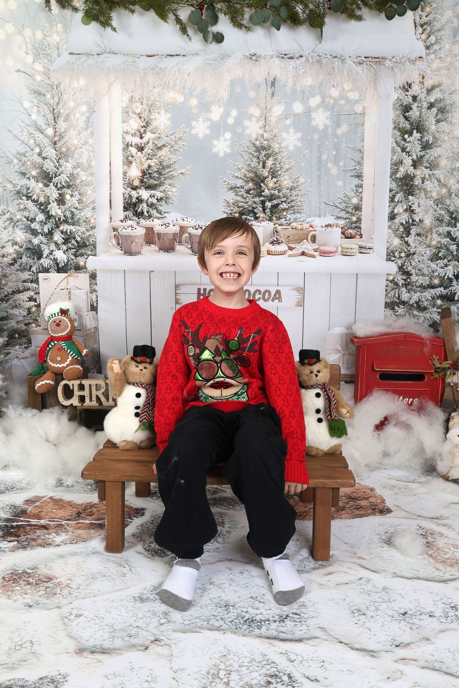 Boy in red reindeer sweater sits on bench amid Christmas decorations, snowmen, and snowy trees for holiday portrait.