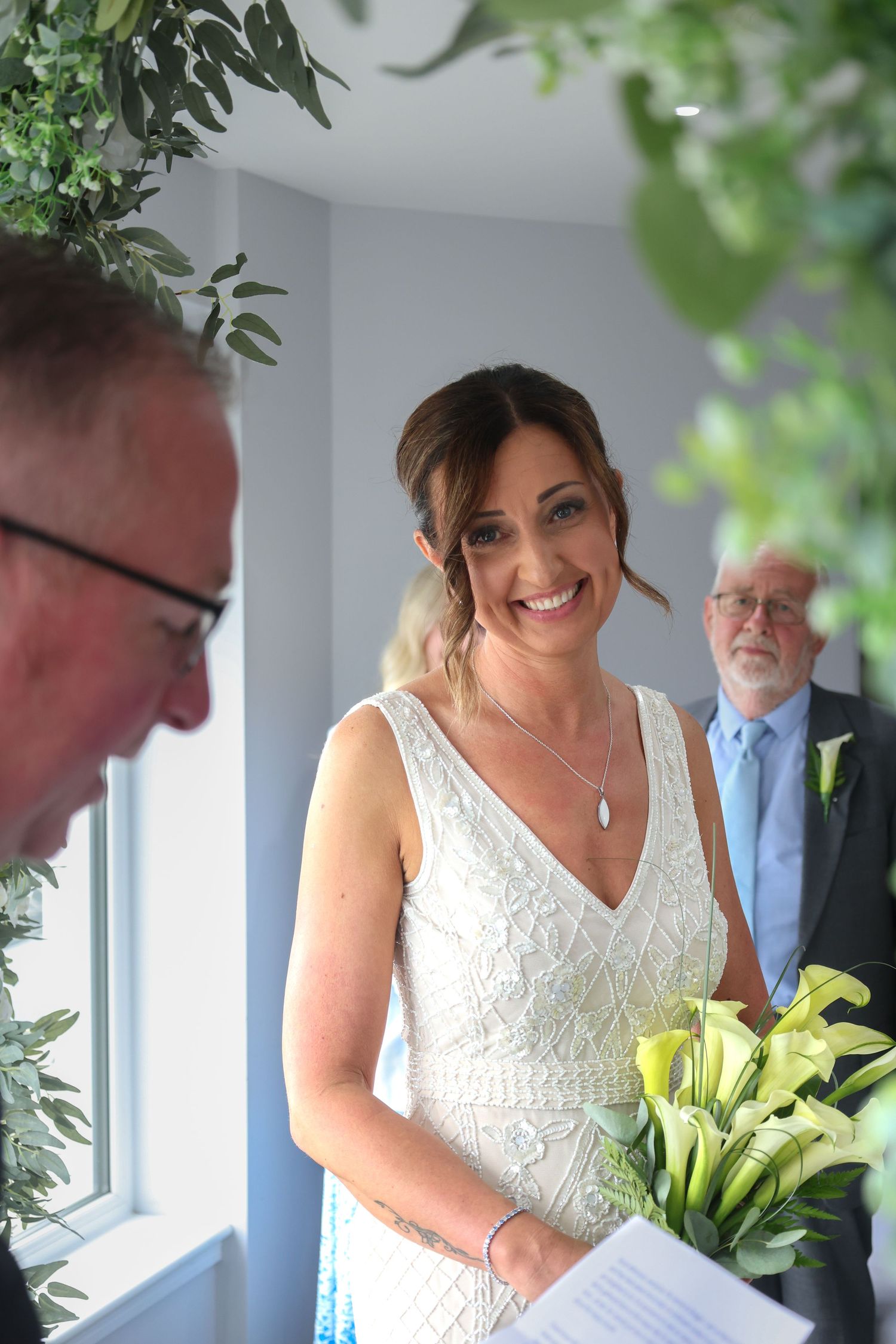 Smiling bride in white lace gown holding calla lily bouquet during wedding ceremony with guests in background.