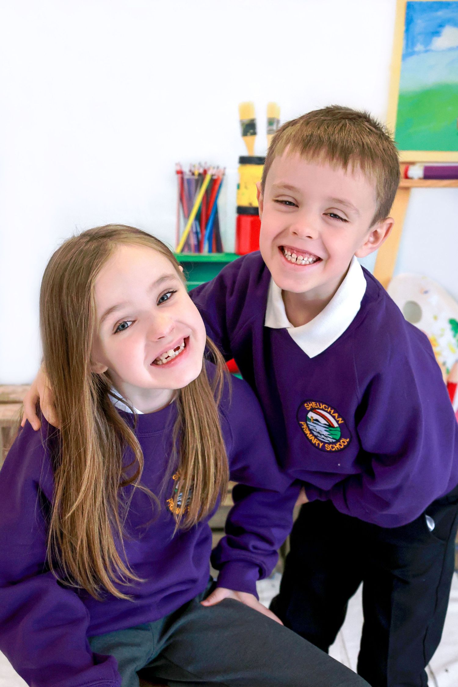 Students in purple school uniforms lean and smile together in a bright classroom setting.