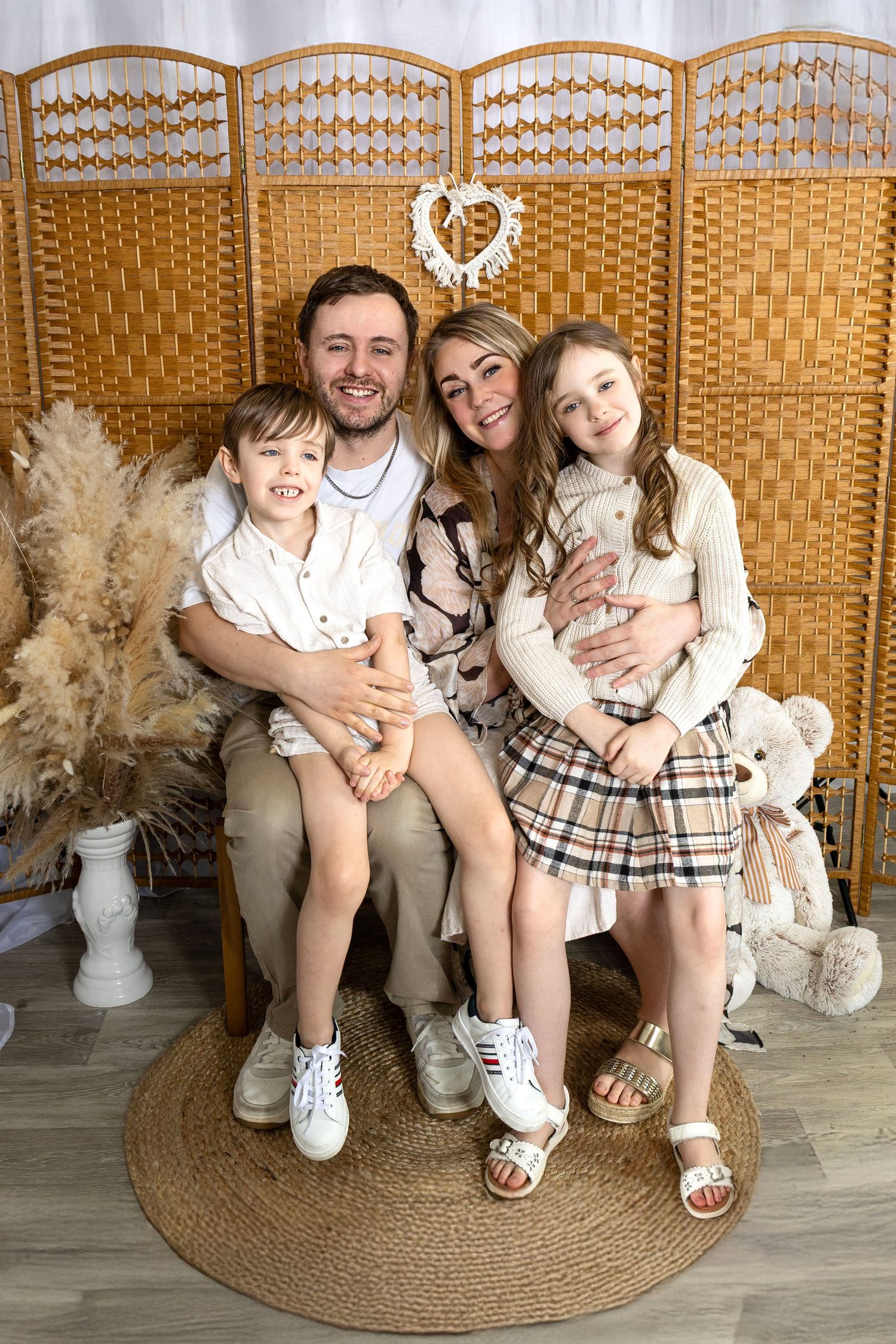 Family sitting together on a round jute rug against a woven room divider with decorative heart and pampas grass decor.