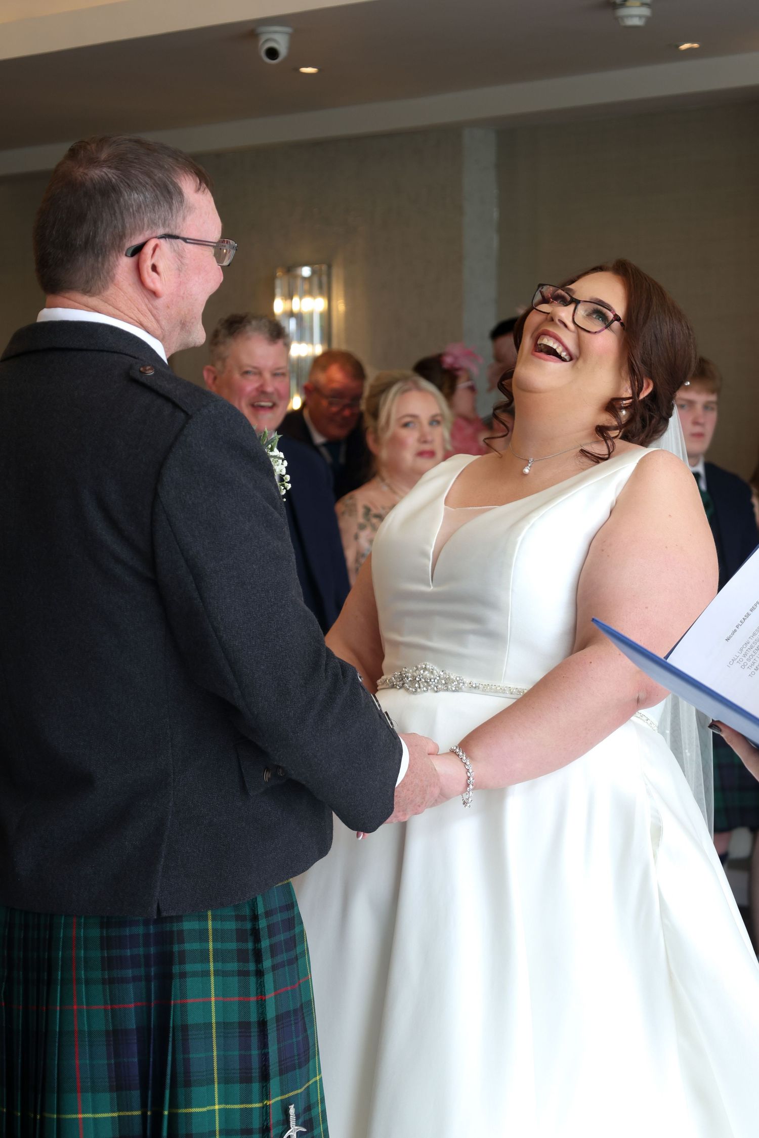Bride in white gown laughing joyfully while holding hands with groom in tartan kilt during wedding ceremony.