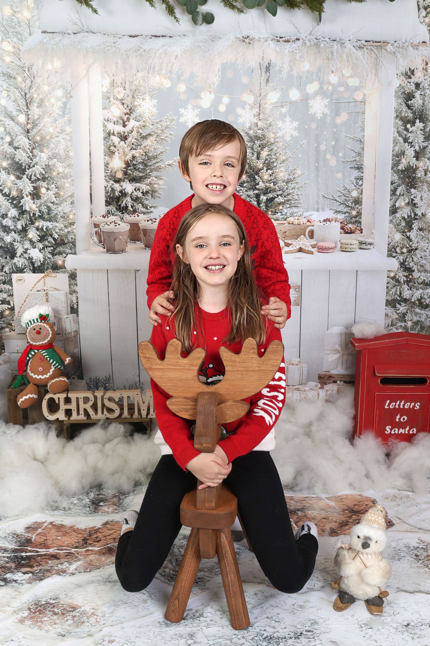 Two children in red Christmas sweaters pose with a wooden reindeer cutout in a festive snowy holiday photo backdrop.