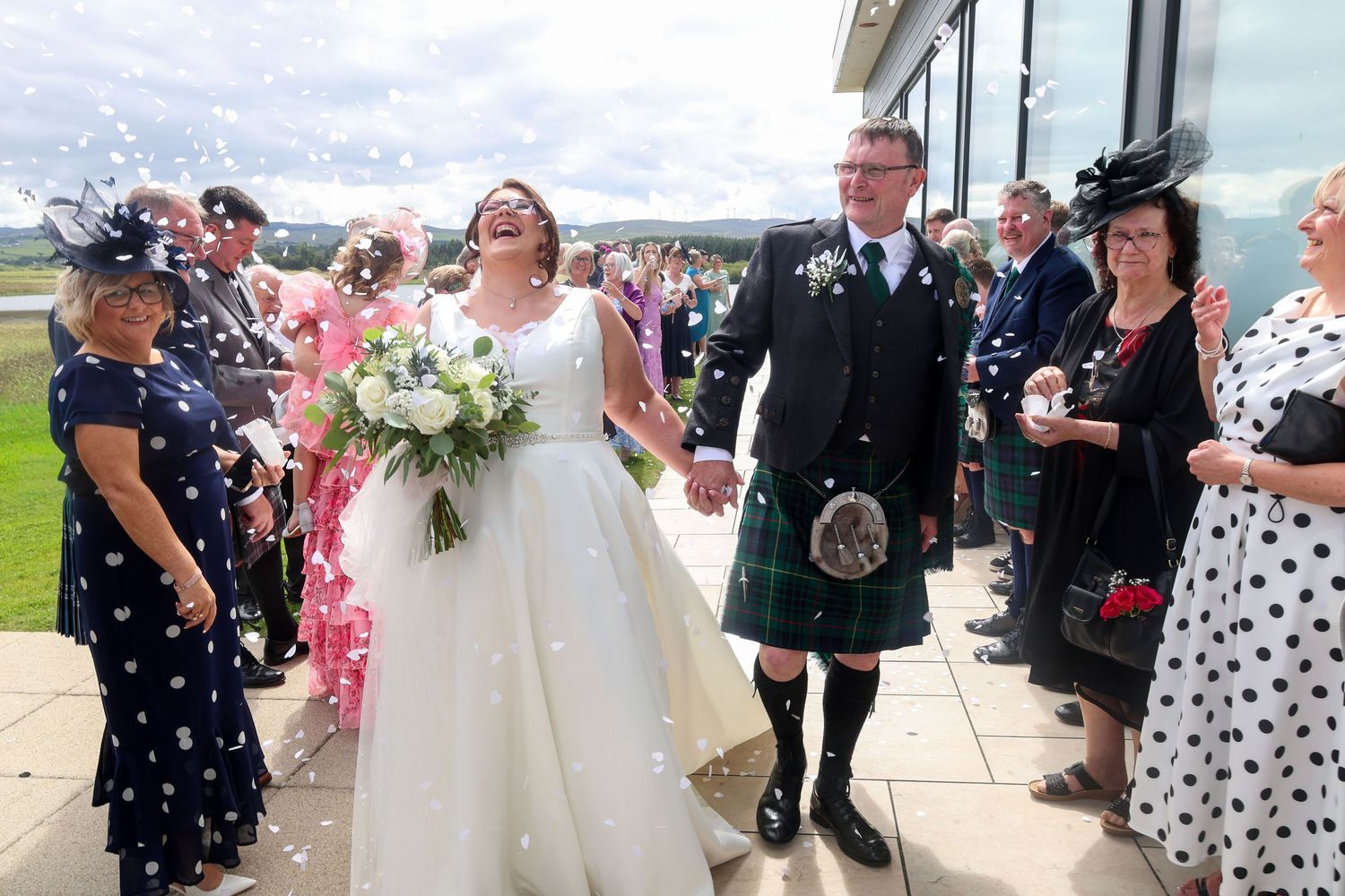 Bride and groom in Scottish kilt celebrate with confetti outside wedding venue surrounded by smiling guests.