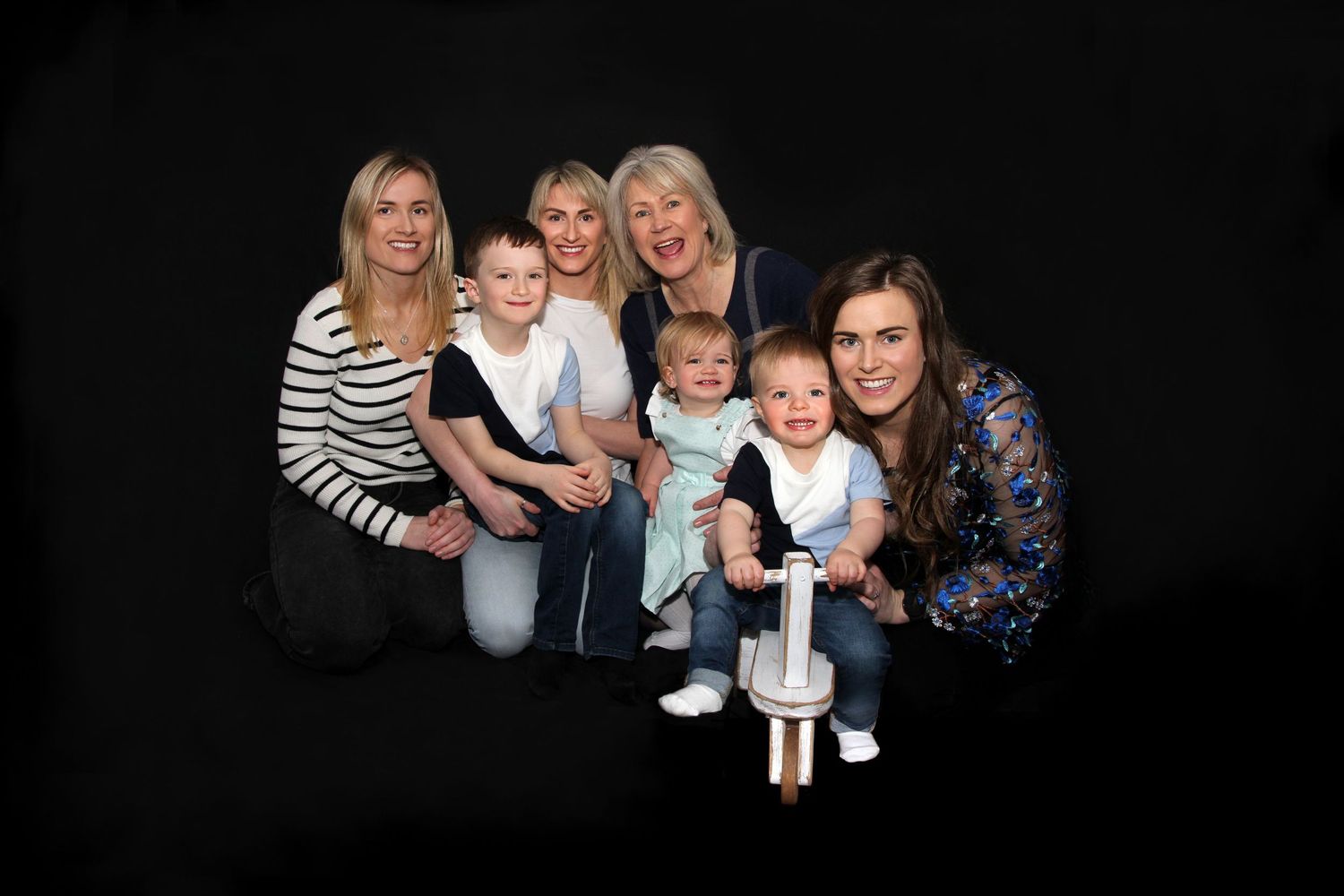 Family portrait of four women with two toddlers posing together against a dark studio background.