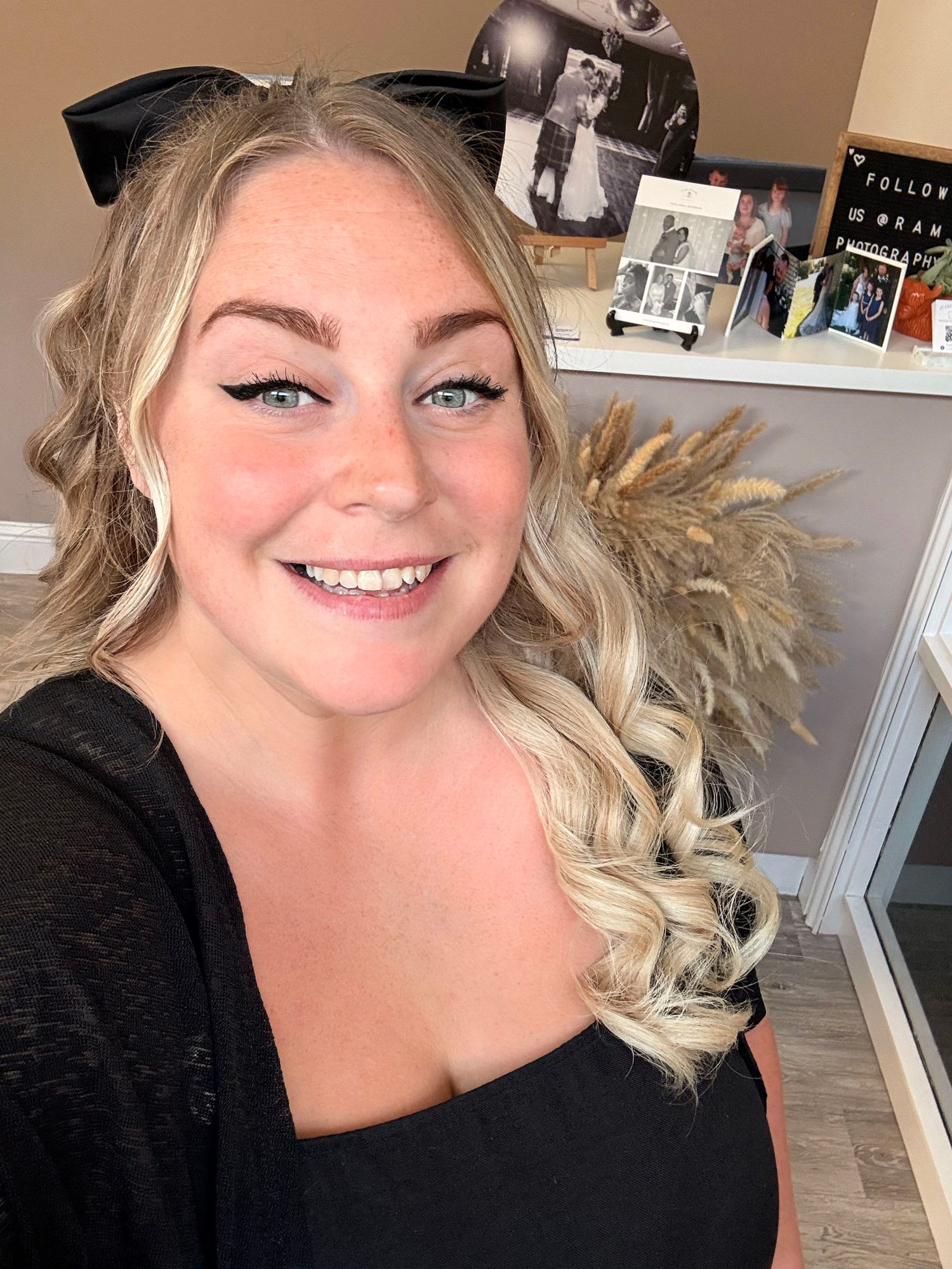 Smiling blonde woman with curly hair and bold eye makeup wearing a black top, taking a selfie indoors.