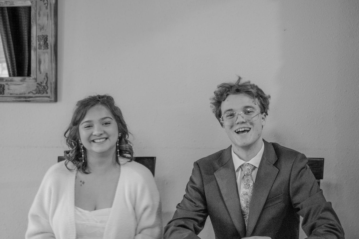 A smiling young couple in formal attire sitting together at a table during a wedding ceremony.