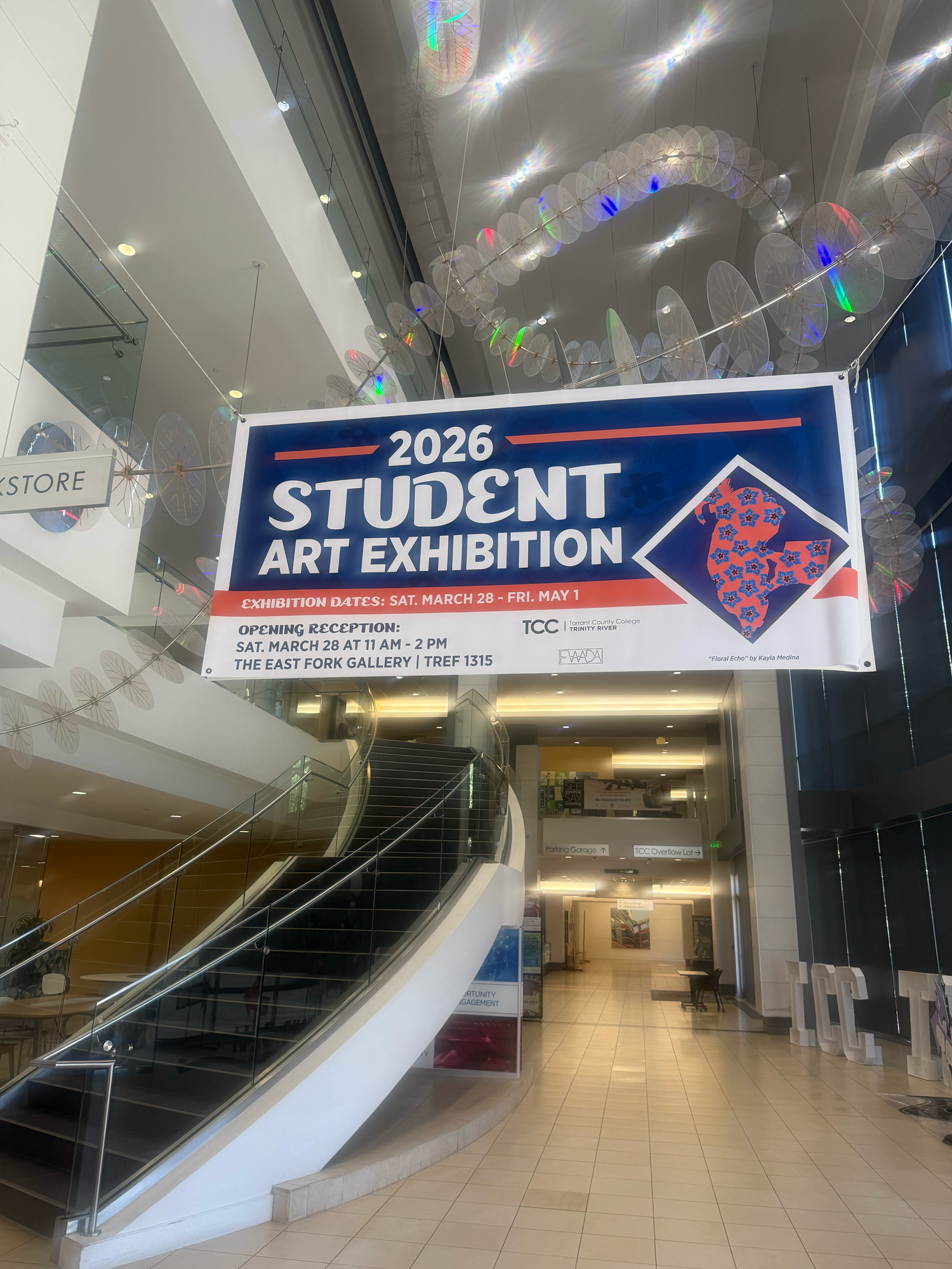 Banner for 2026 Student Art Exhibition at TCC displayed above an escalator in a modern building.
