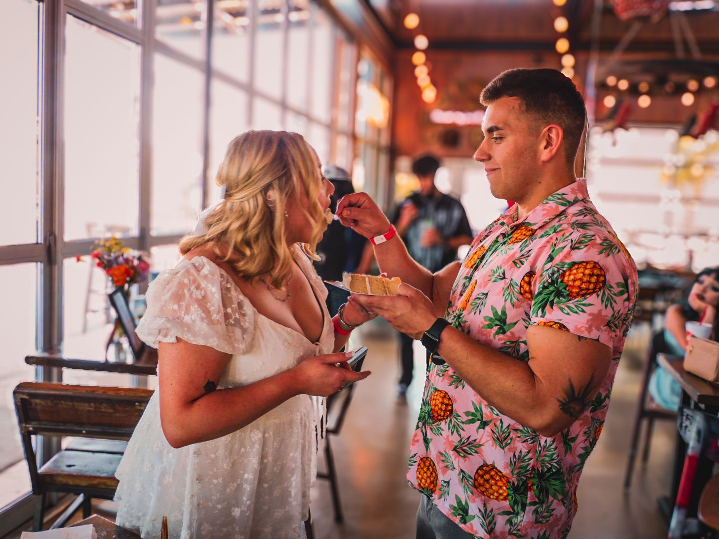 A couple in casual wedding attire share a drink at a festive indoor reception venue with string lights.