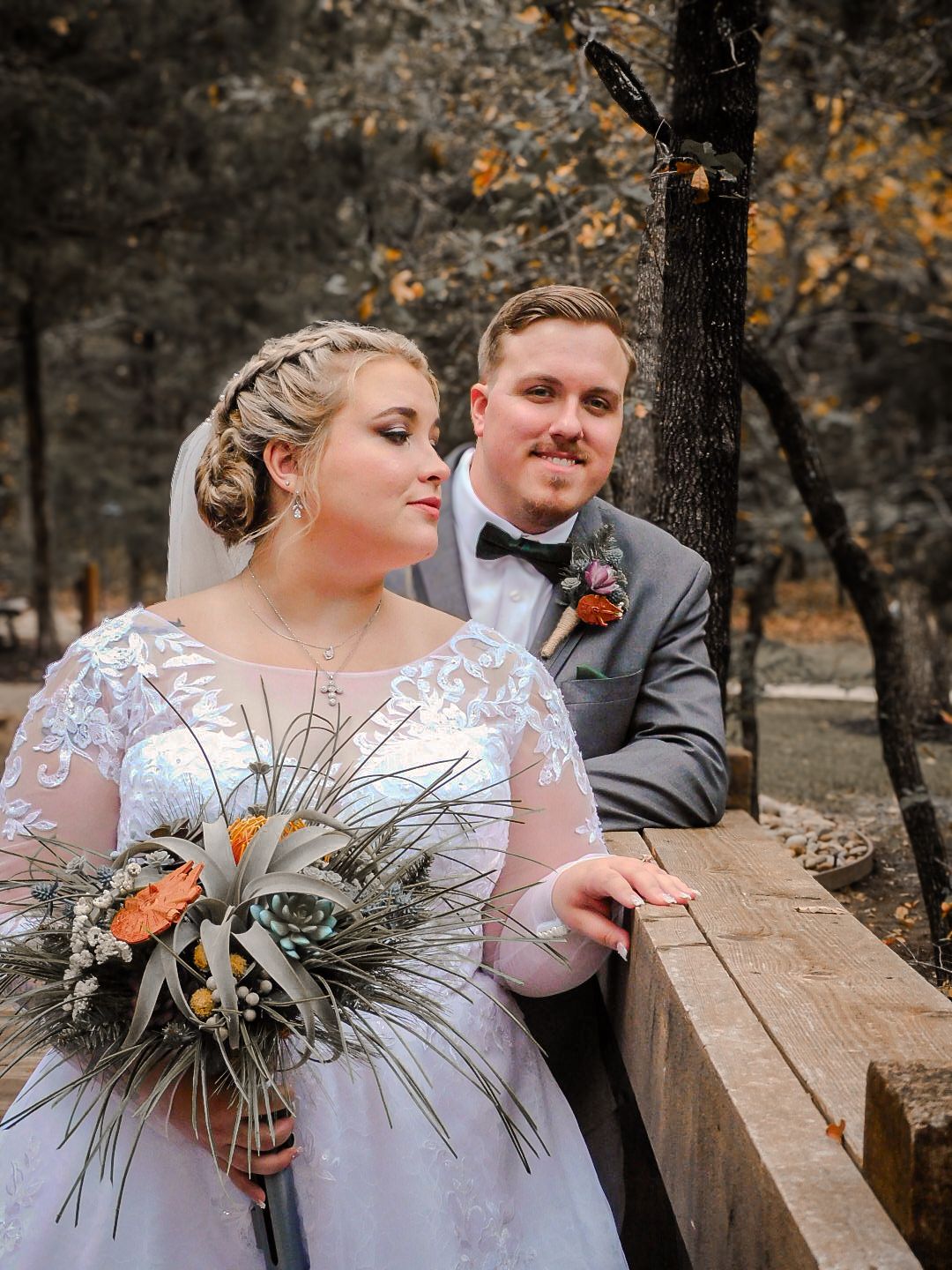 A bride holding a rustic bouquet and groom in gray suit pose outdoors in a fall forest setting.