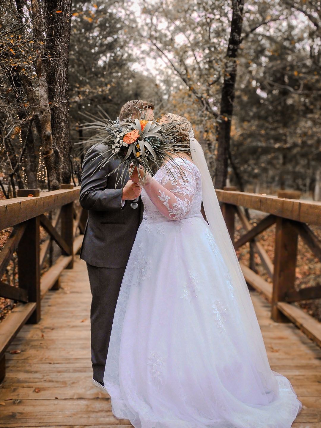 A bride and groom share a kiss hidden behind a bouquet on a wooden bridge surrounded by autumn trees.