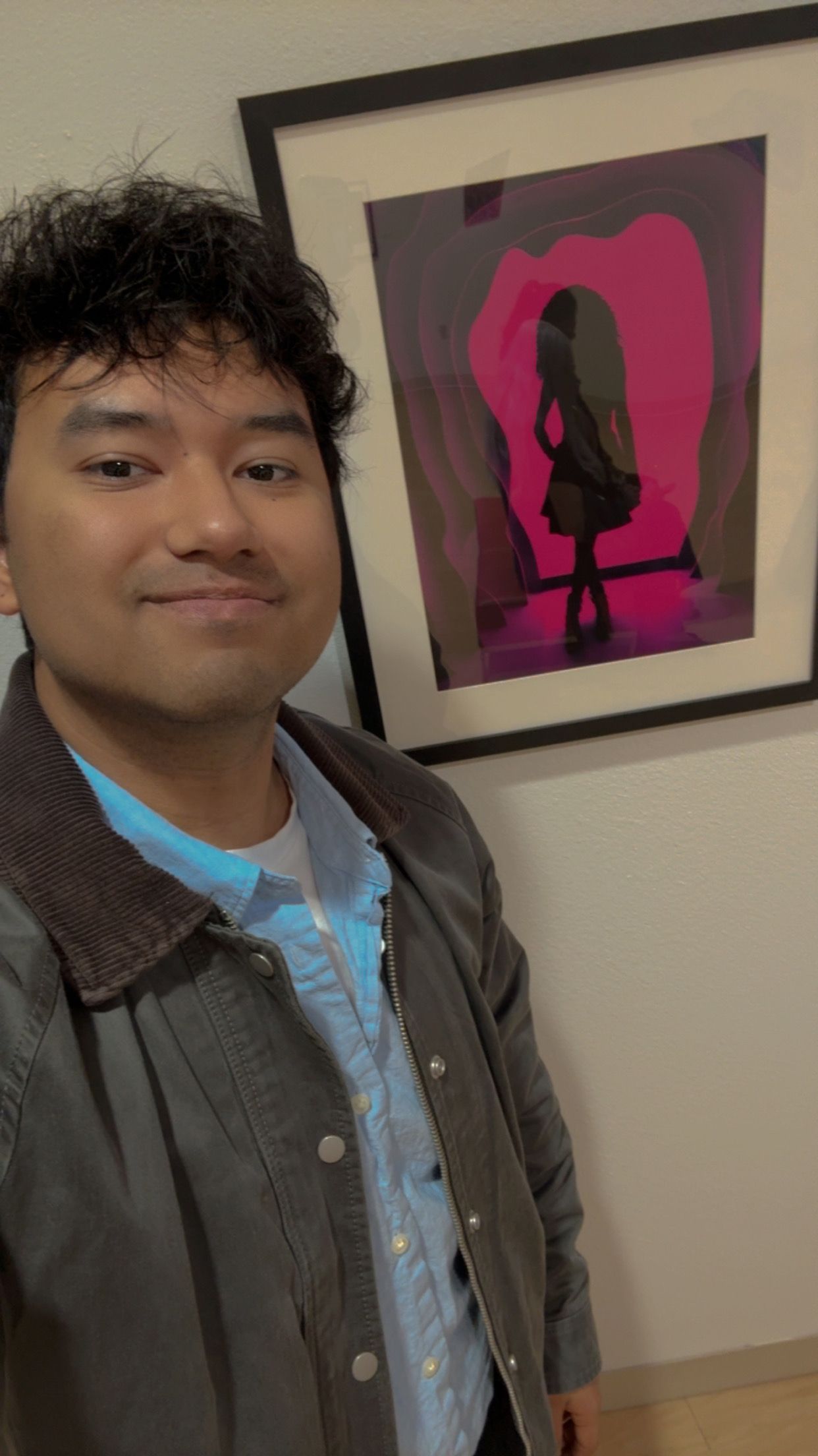 Young man smiles in front of a framed pink silhouette artwork at an art gallery reception.