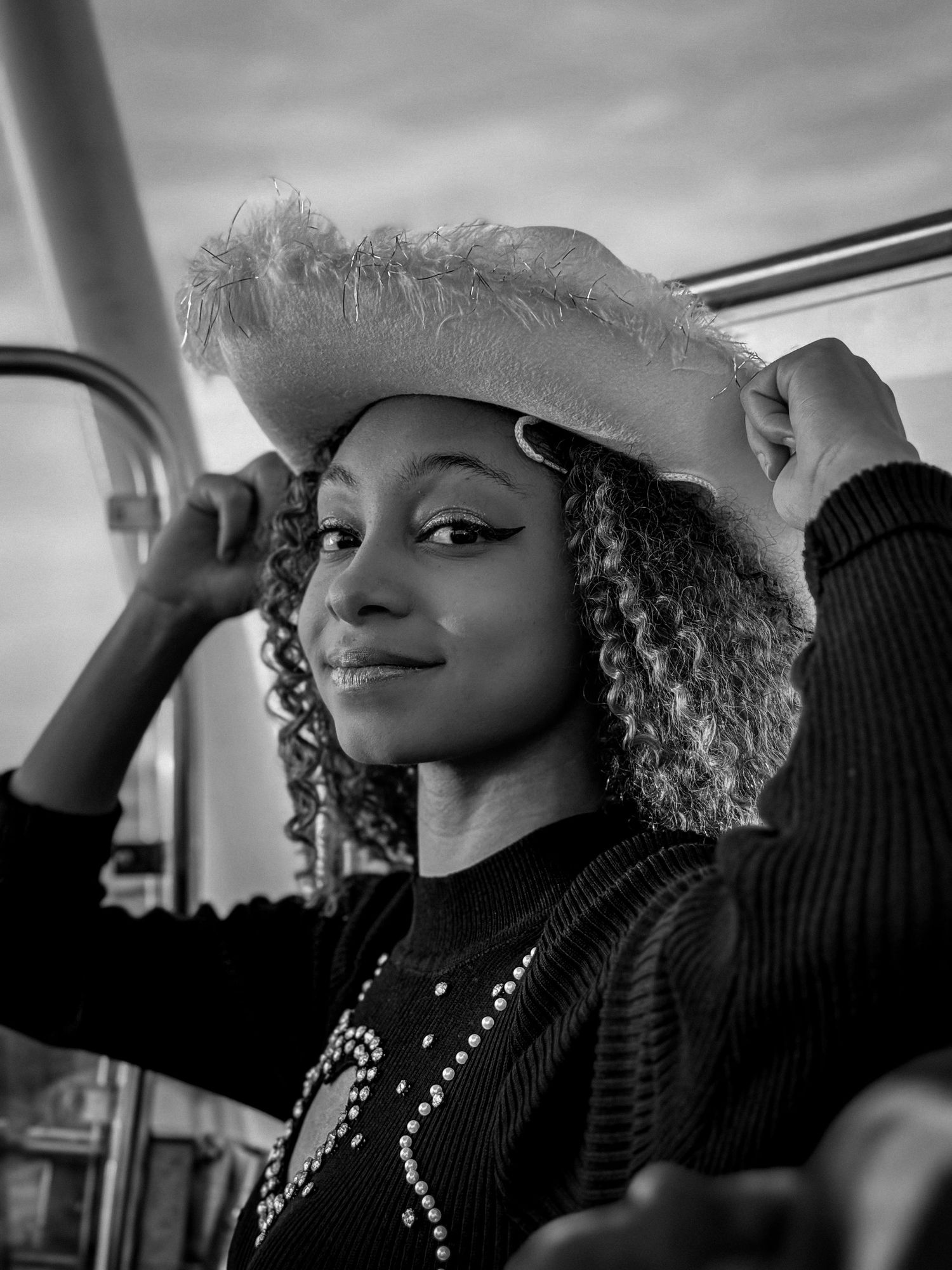 Young woman with curly hair smiles wearing a fuzzy white hat in a black and white portrait.
