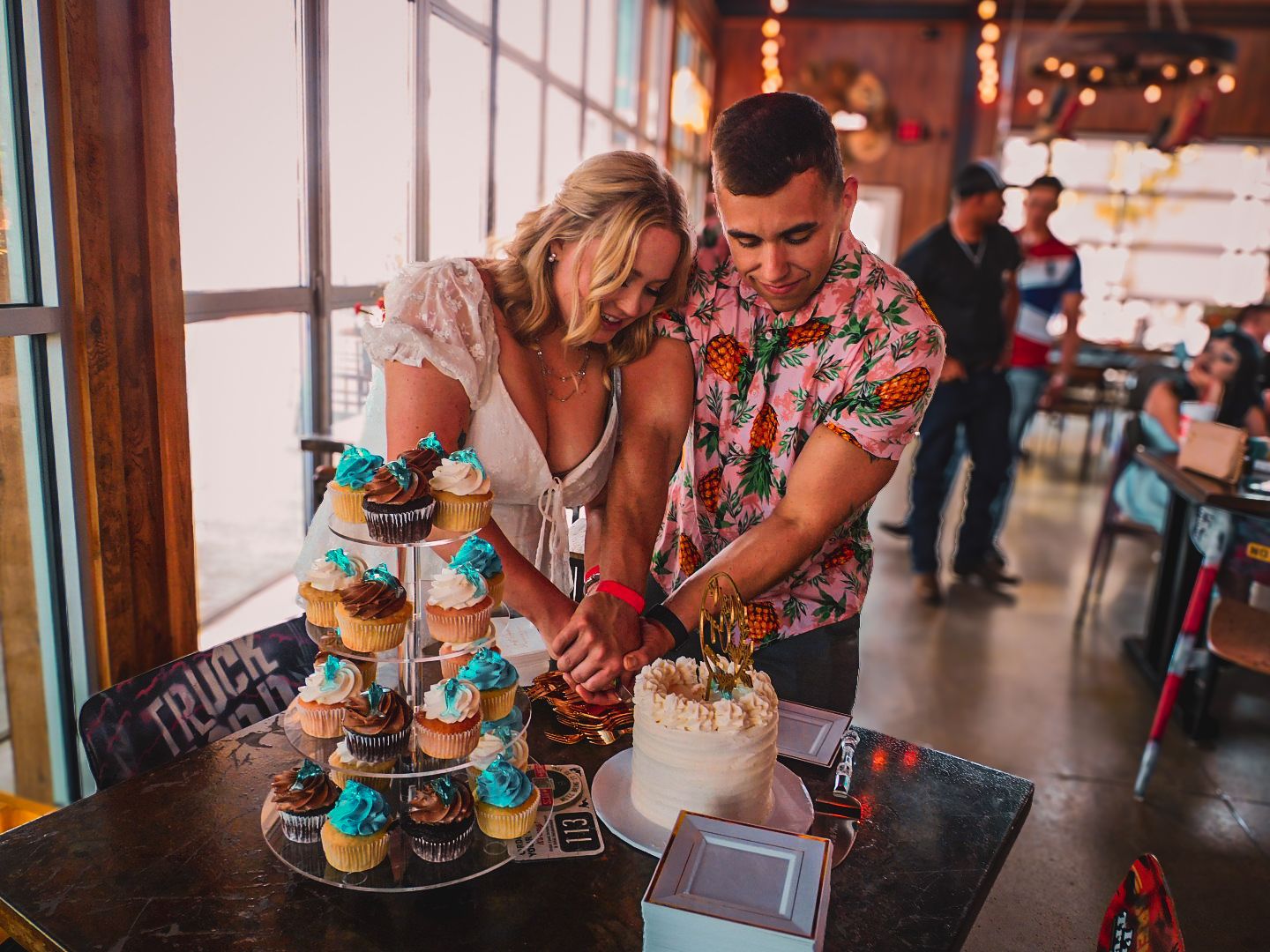 A couple cuts their wedding cake surrounded by colorful cupcakes at a casual tropical-themed reception.