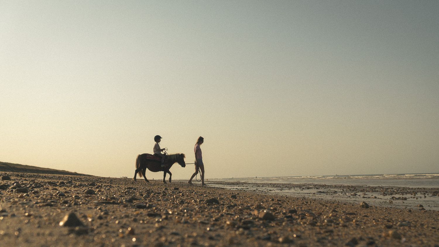 Two silhouetted figures with a donkey walking along a sandy beach at dusk.
