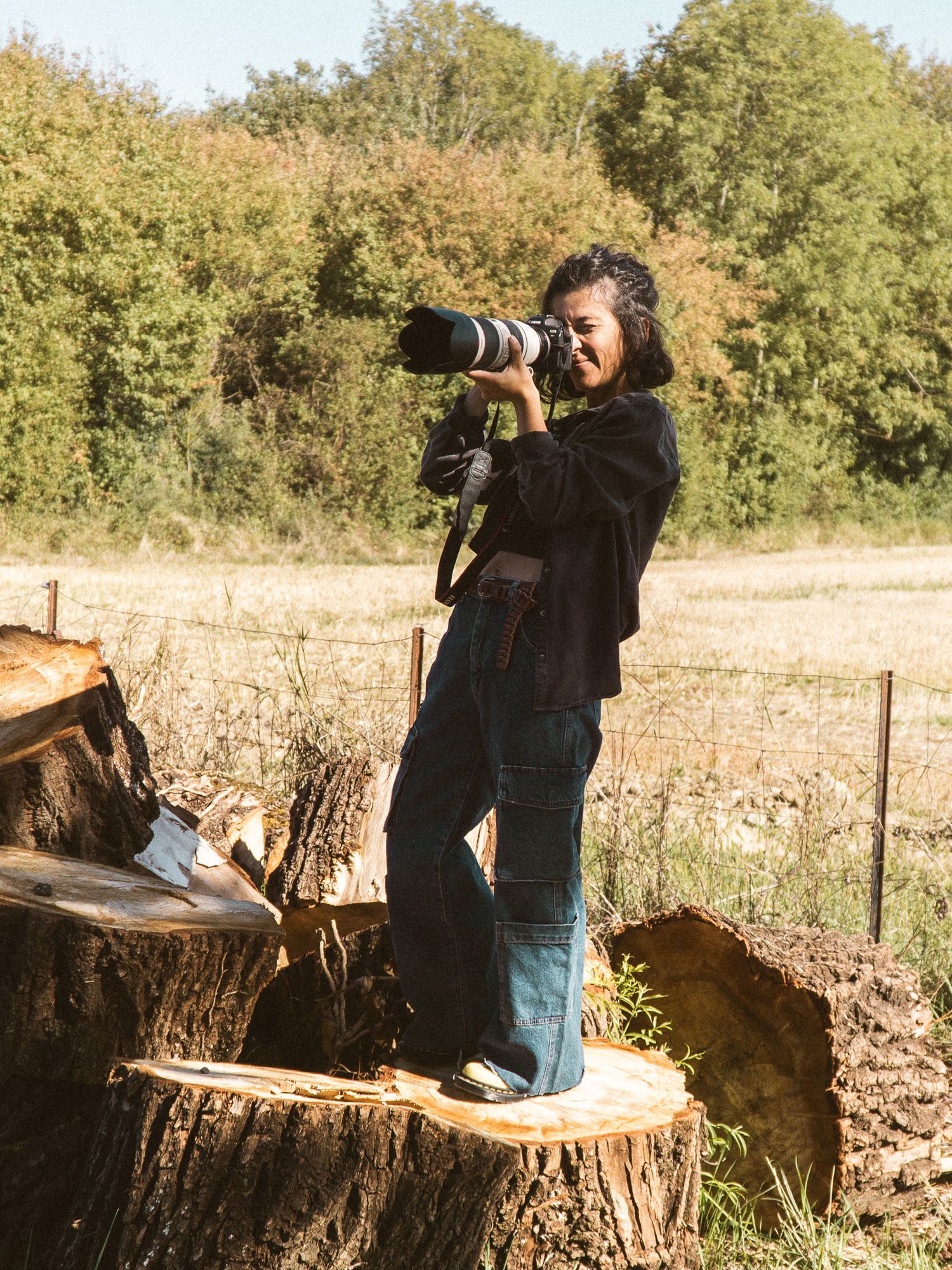 Female photographer standing on a tree stump using a telephoto lens camera in a sunny field.