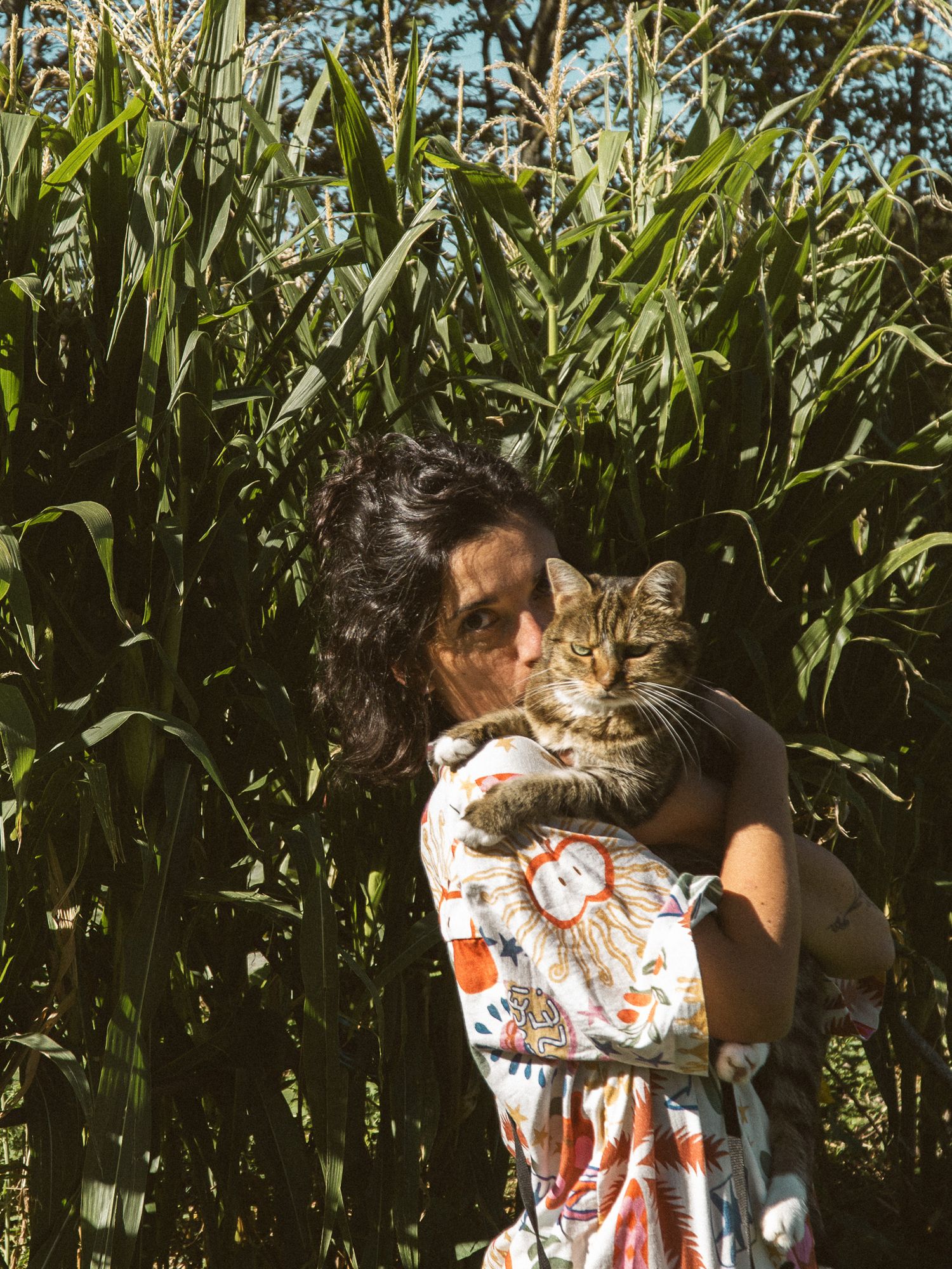 Woman holding a tabby cat in front of a lush green cornfield on a sunny day.
