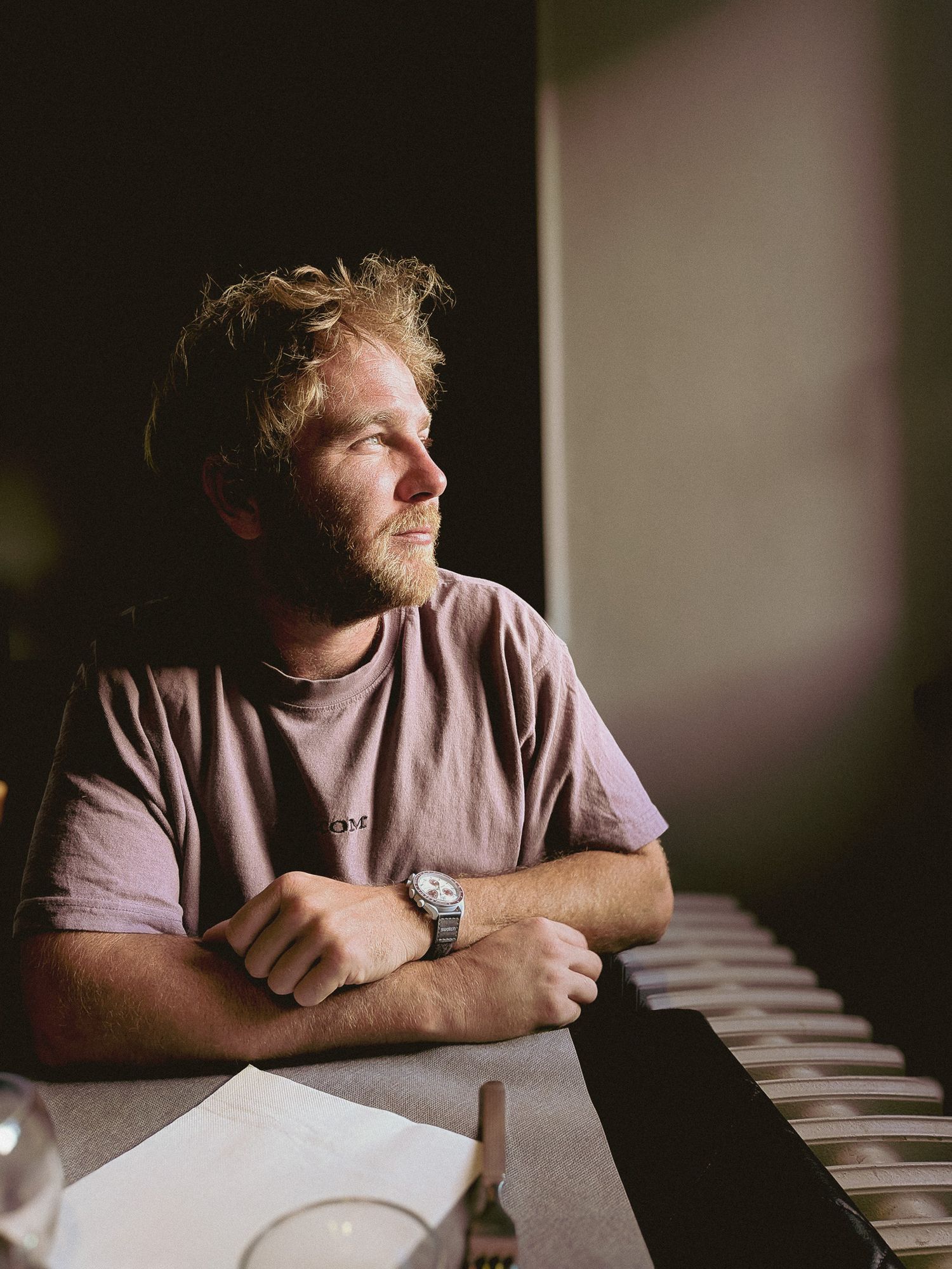 A young man with curly blonde hair and beard sits at a table, wearing a pink t-shirt and watch, gazing thoughtfully.