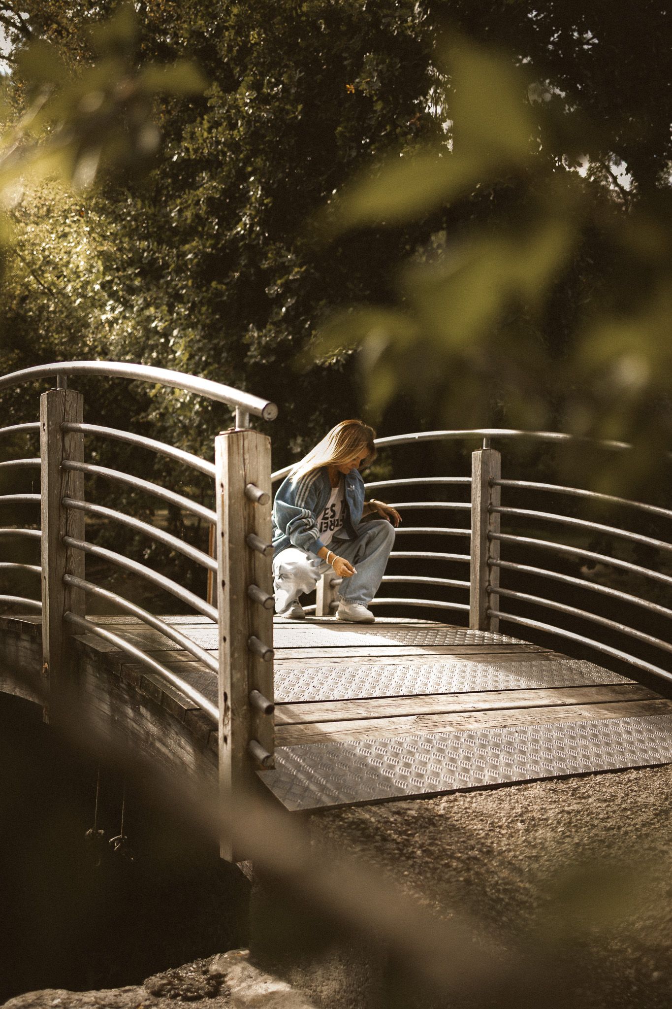 A young woman crouches on a metal footbridge surrounded by lush green trees in warm sunlight.