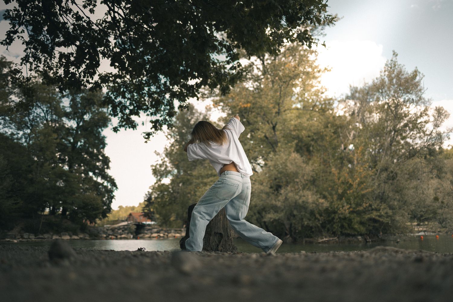 Person dancing freely under large trees in a sunlit outdoor natural setting.