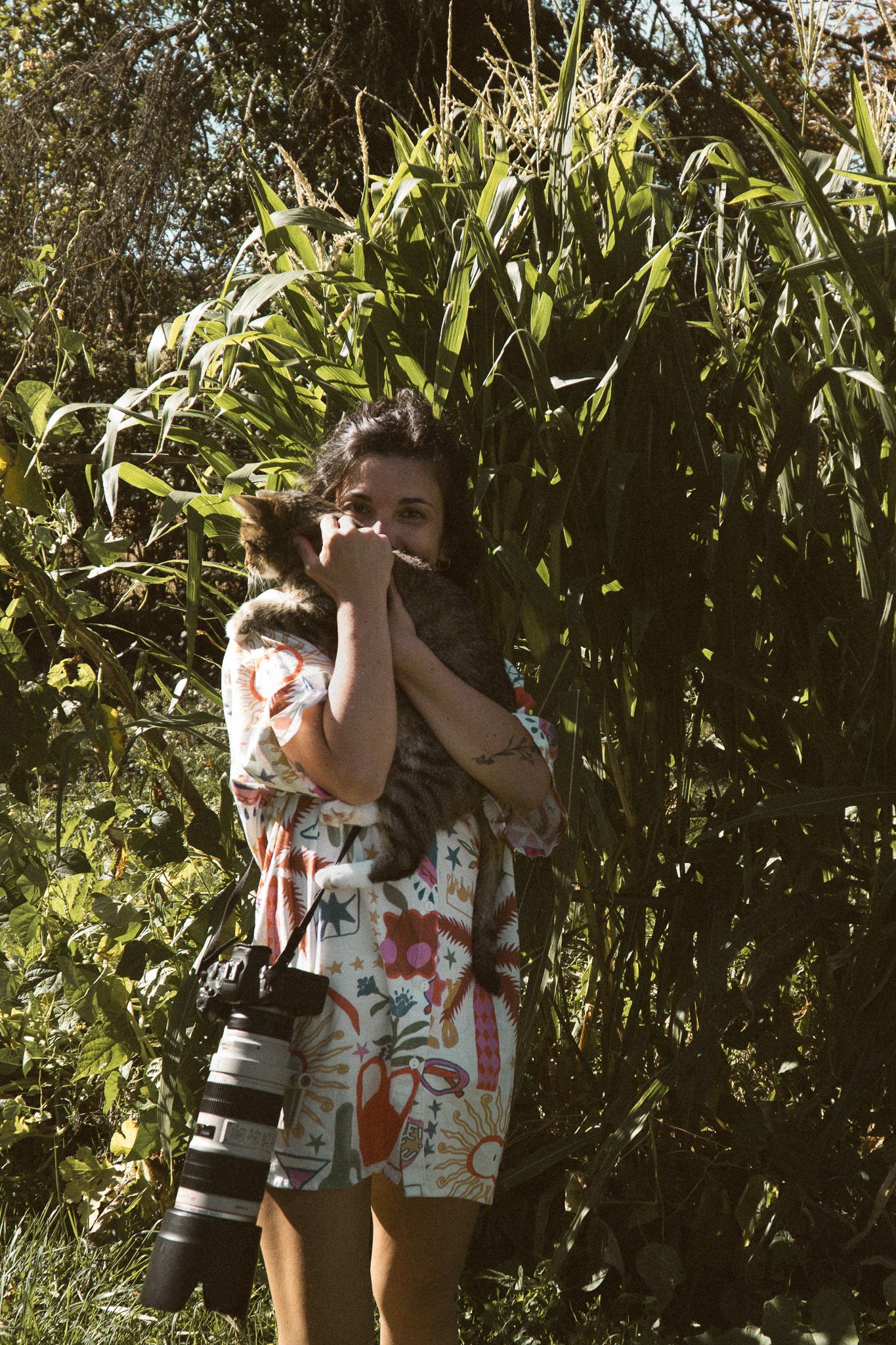 Woman in floral outfit holds a cat while carrying a camera lens outdoors among lush green foliage.