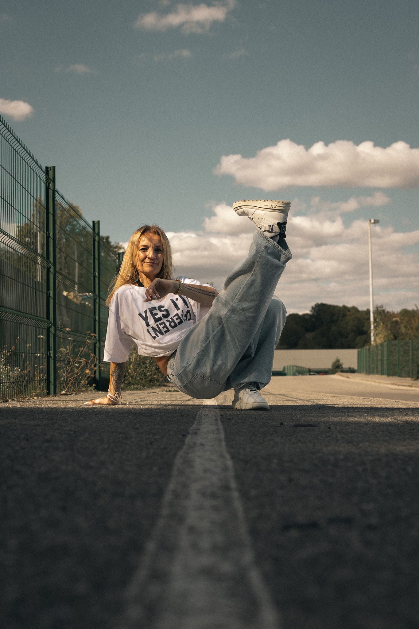A blonde woman in a white graphic tee performs a floor dance move on an outdoor court.