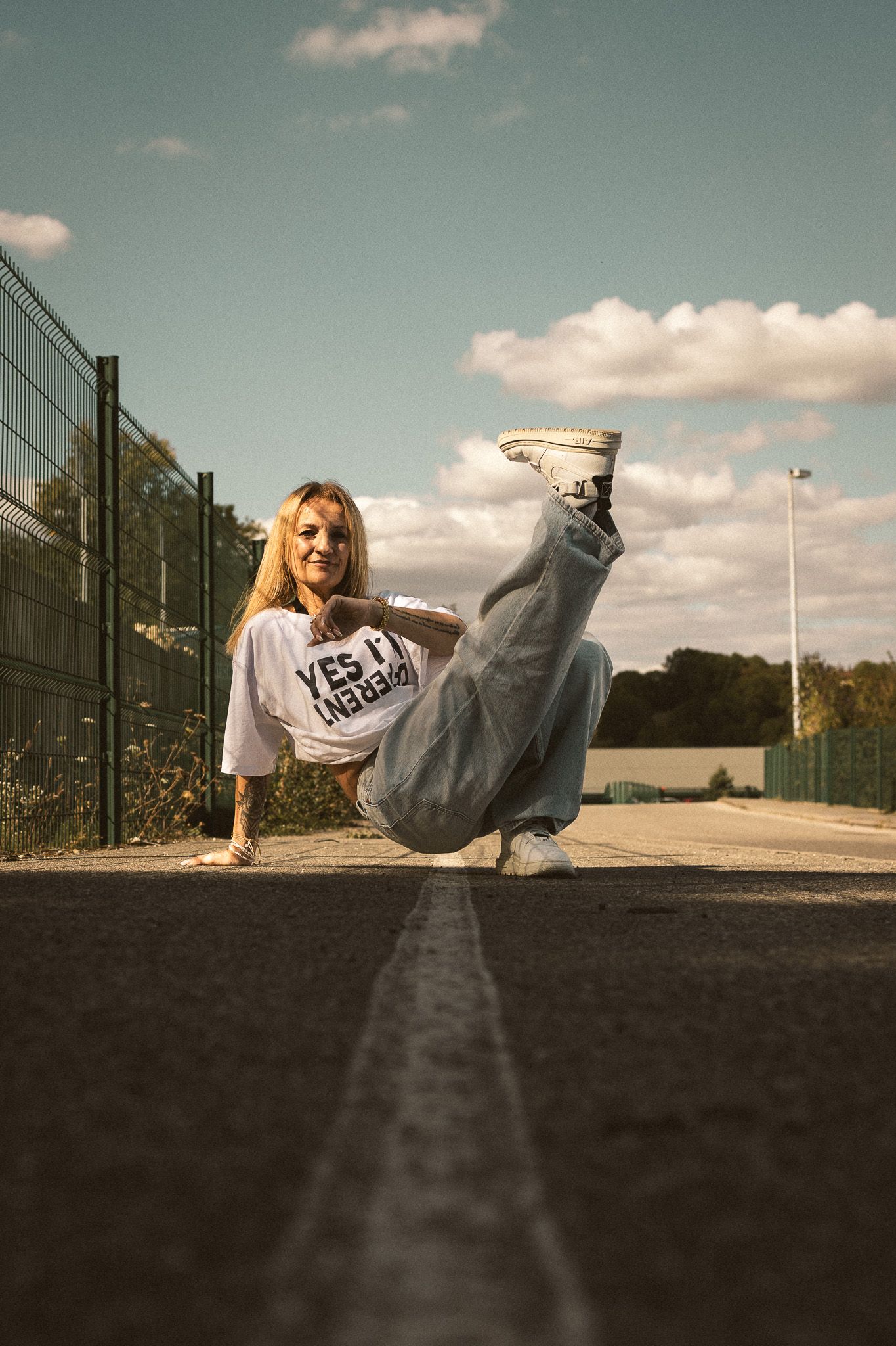 Blonde woman performing a breakdance freeze move on an outdoor court under a blue cloudy sky.